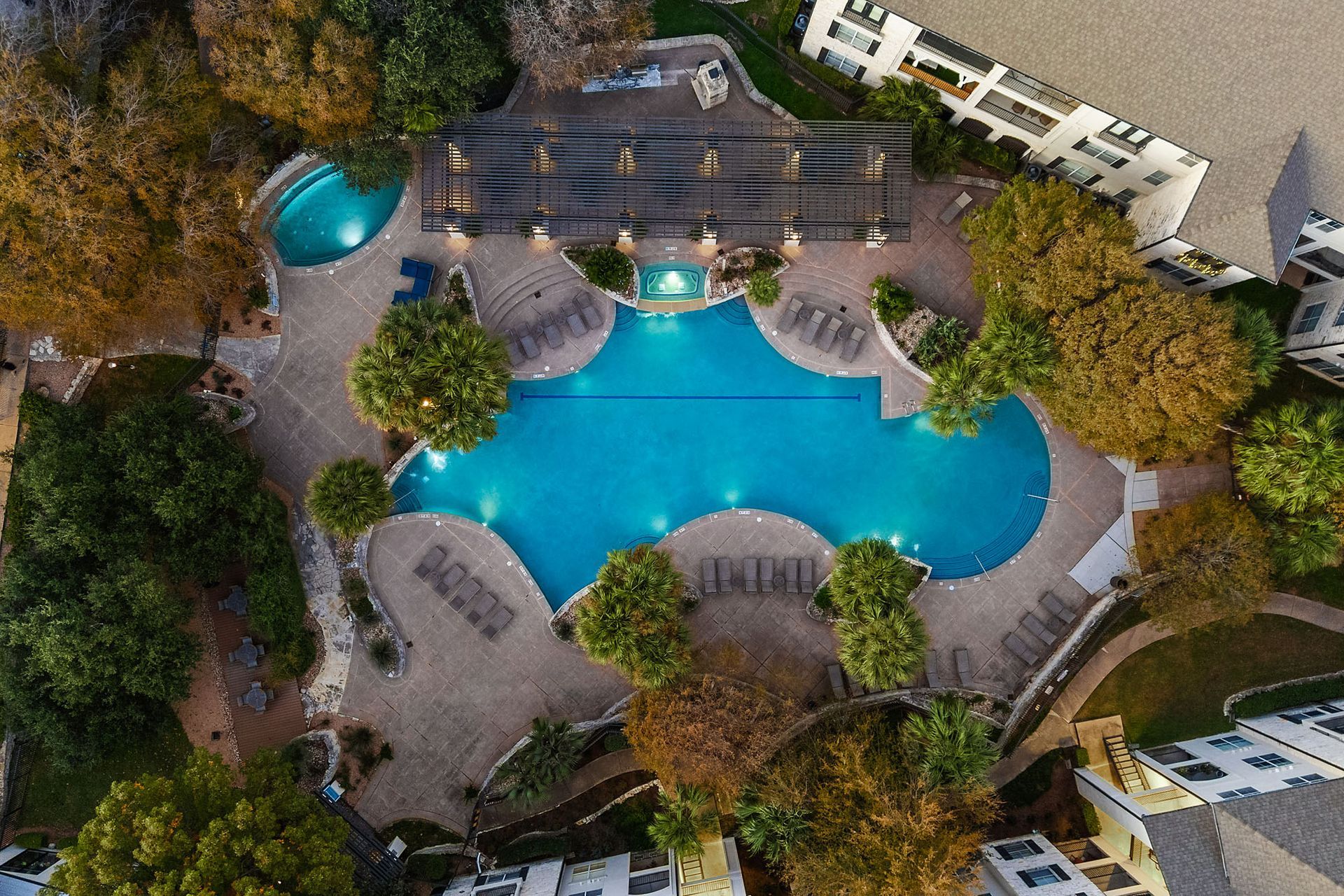 Aerial view of a bright blue swimming pool with lounge chairs and trees in a landscaped courtyard at Marquis at Great Hills in Austin, TX.