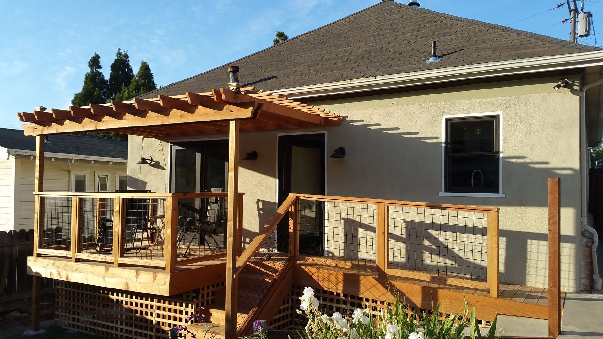 Backyard deck with wooden pergola and railing attached to a beige house in sunlight