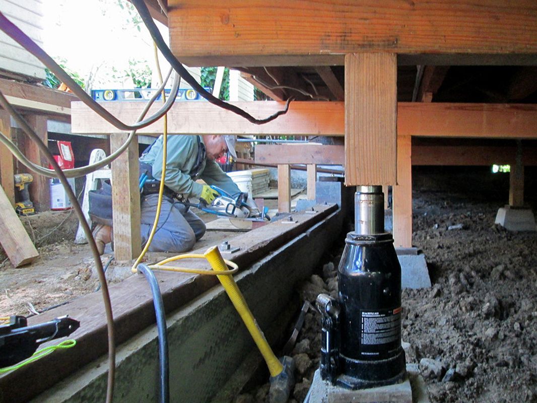 A man is working under a wooden structure with a hydraulic jack in the foreground