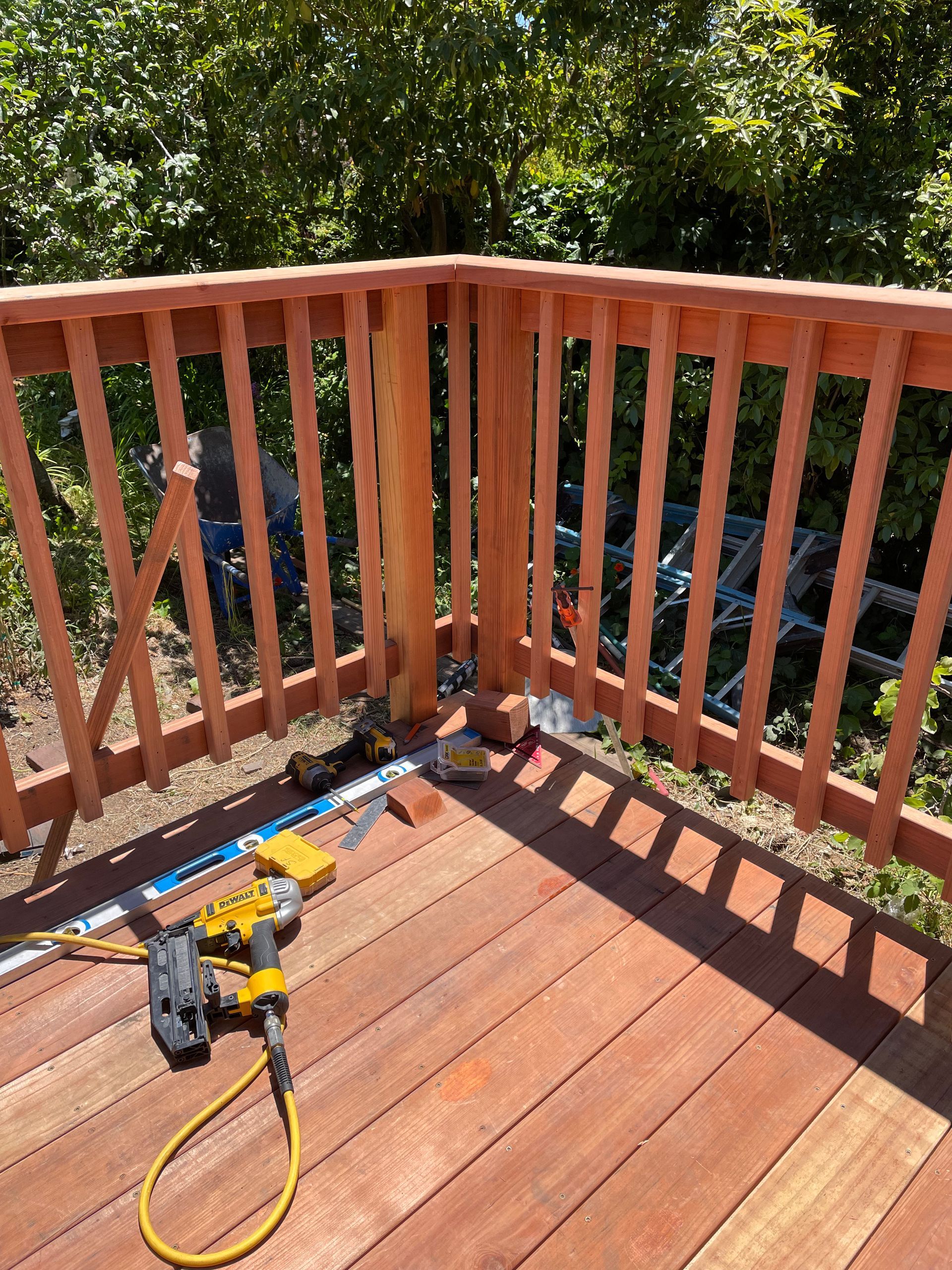 Wooden deck corner with red railing, power tools, and a yellow hose in bright sunlight