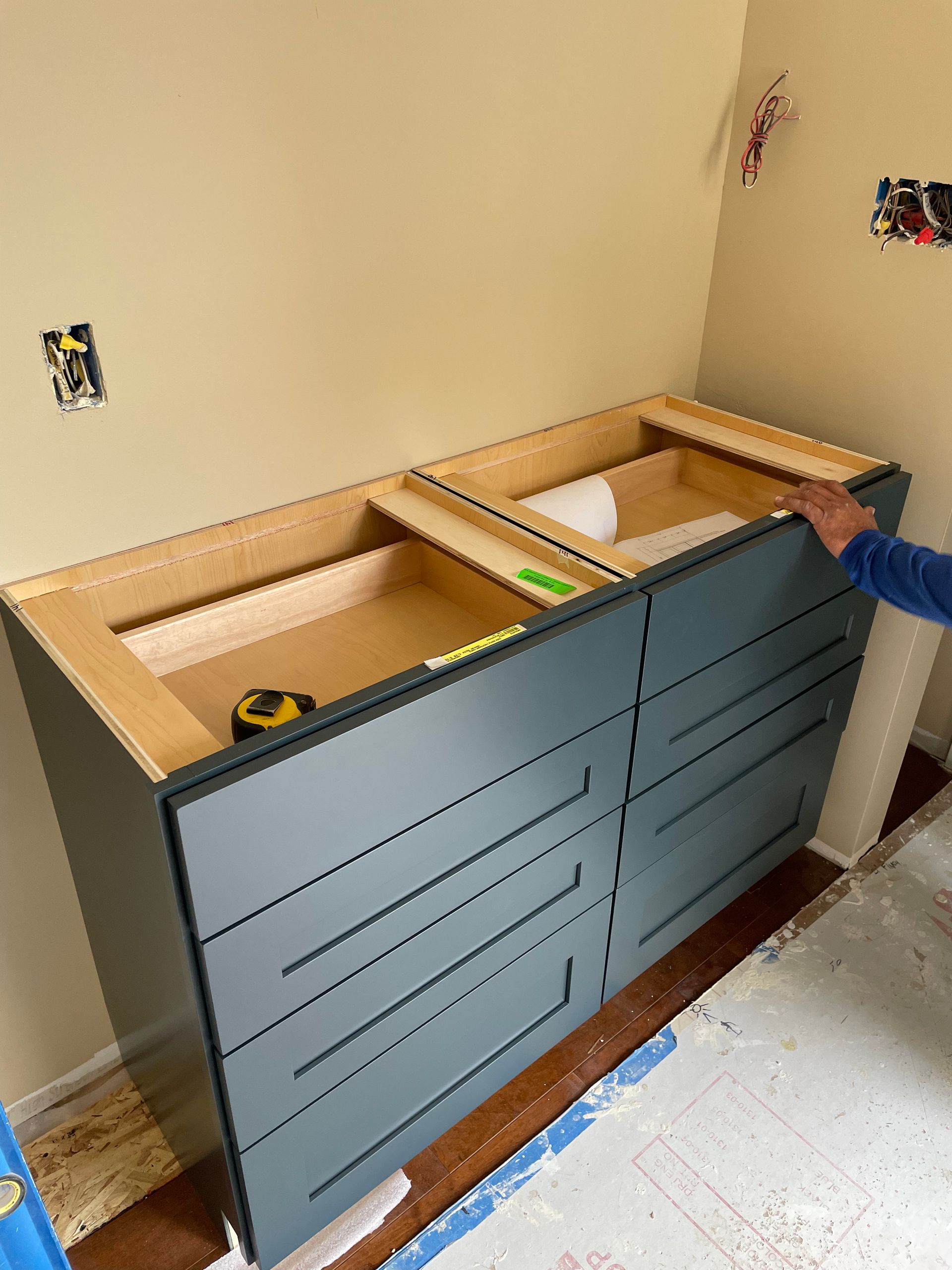 Blue vanity cabinet with open countertop cutouts under construction in a beige room, measuring tape inside.