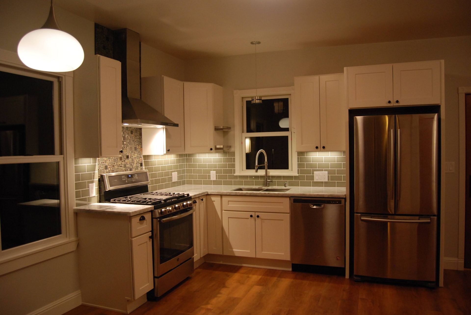 Modern kitchen with white cabinets, stainless steel appliances, and warm hardwood floors at night