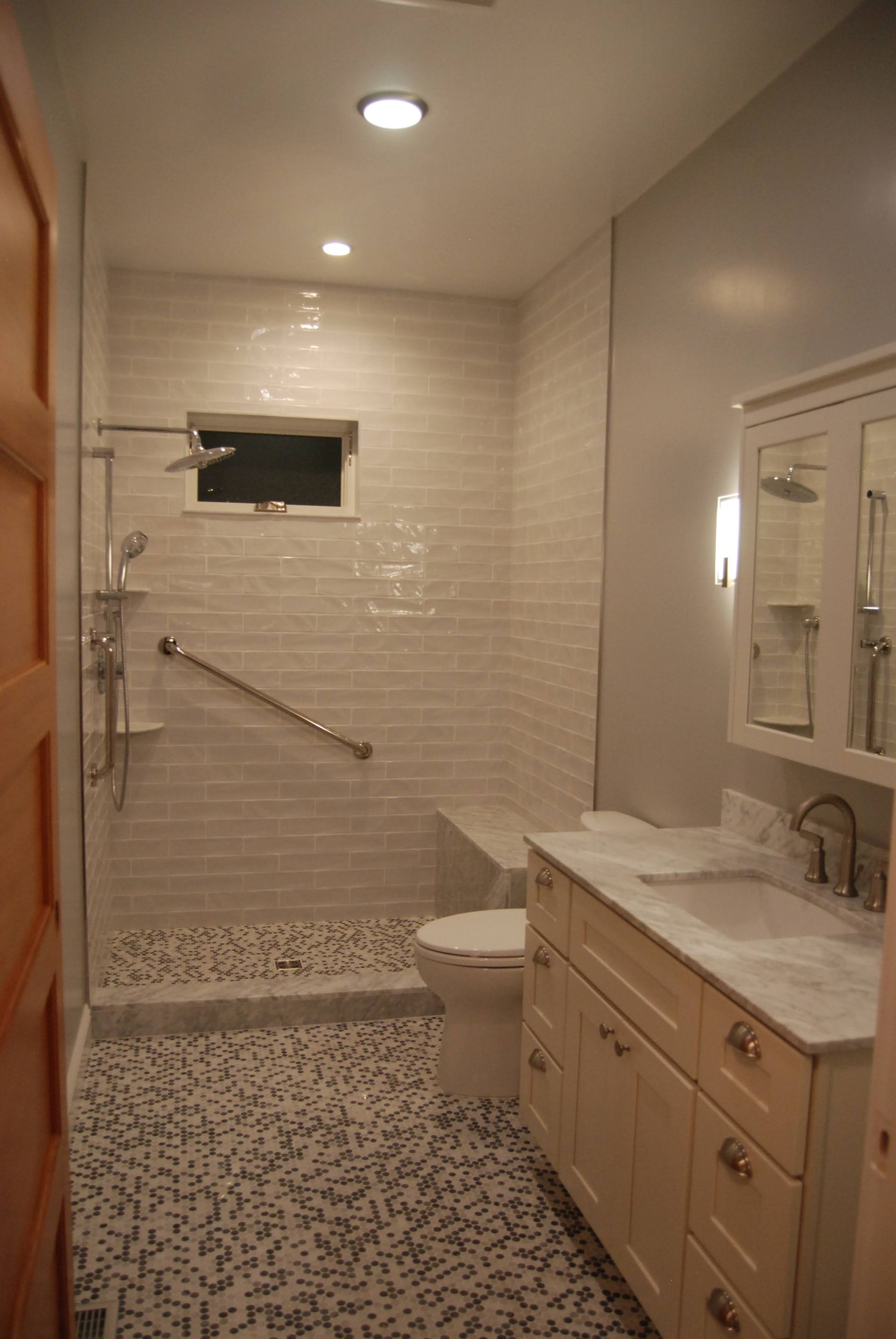 Narrow accessible bathroom with tiled walk-in shower, grab bar, toilet, and vanity under bright lights