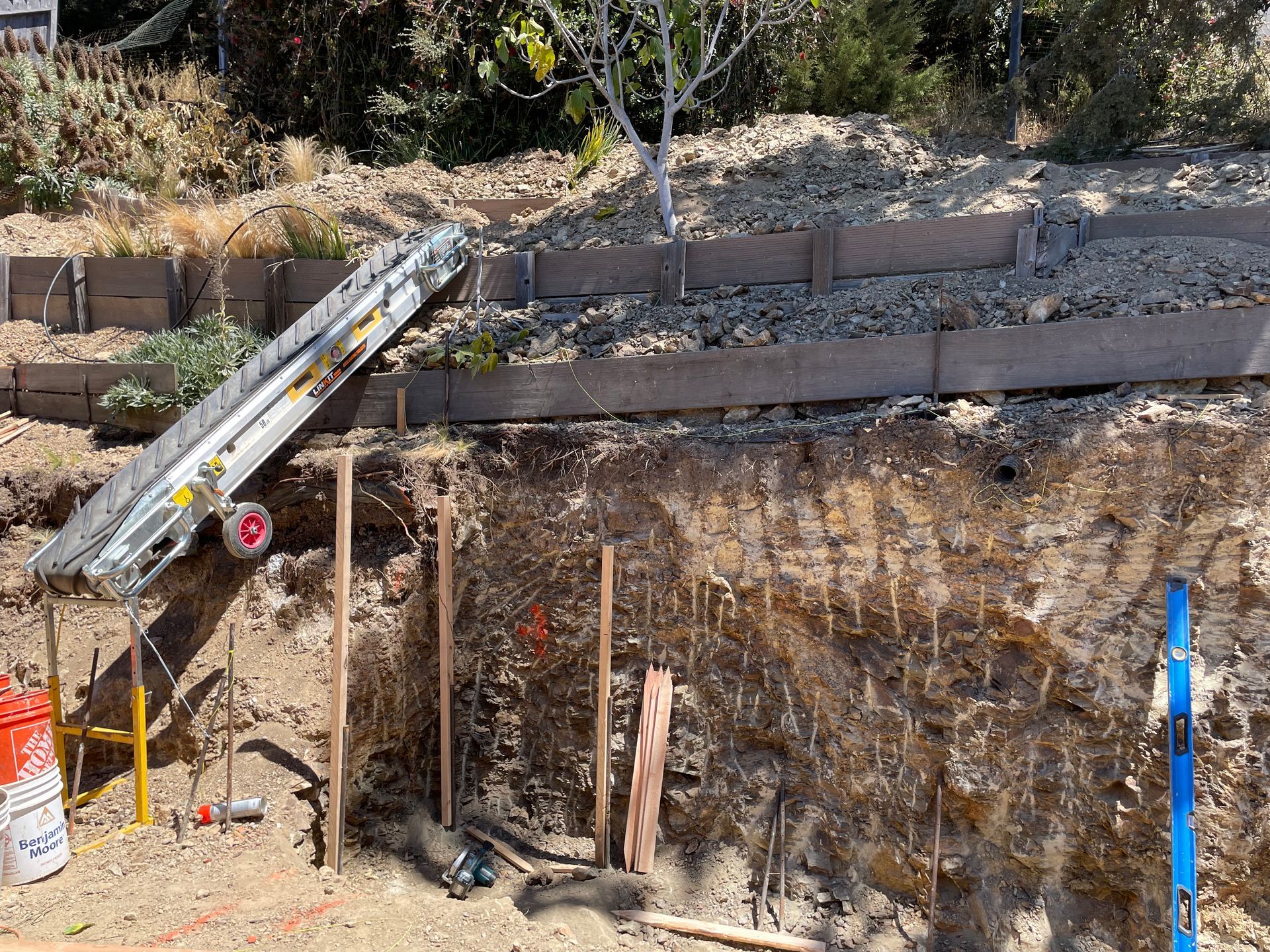 Construction site excavation with retaining wall, ladder, and digging tools beside a dirt embankment