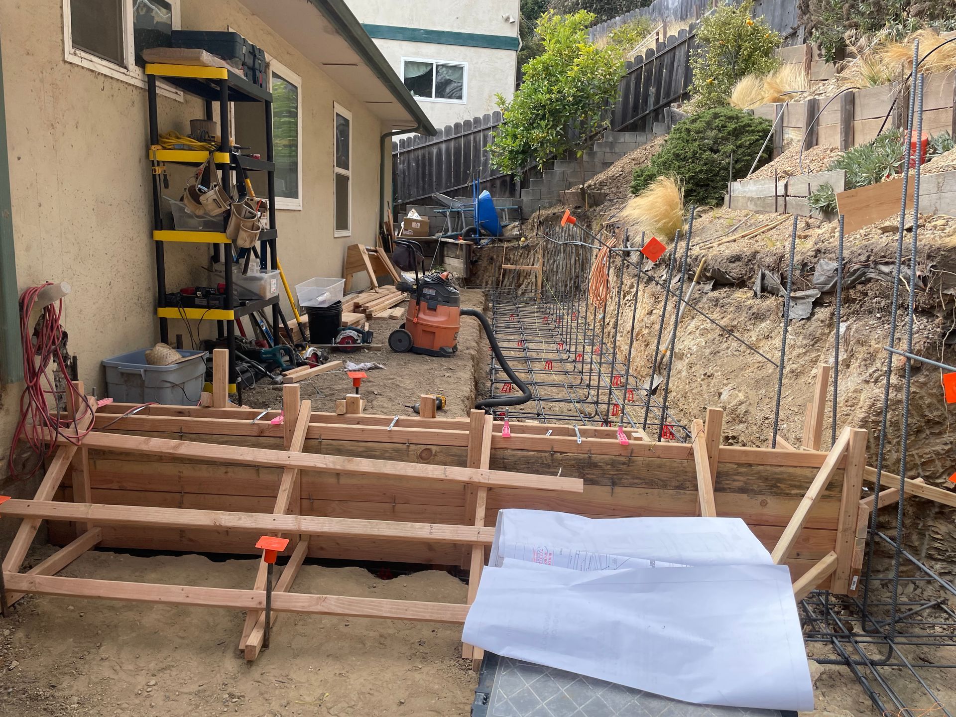 Construction site with wooden formwork, rebar, tools, and a partially covered trench beside a house