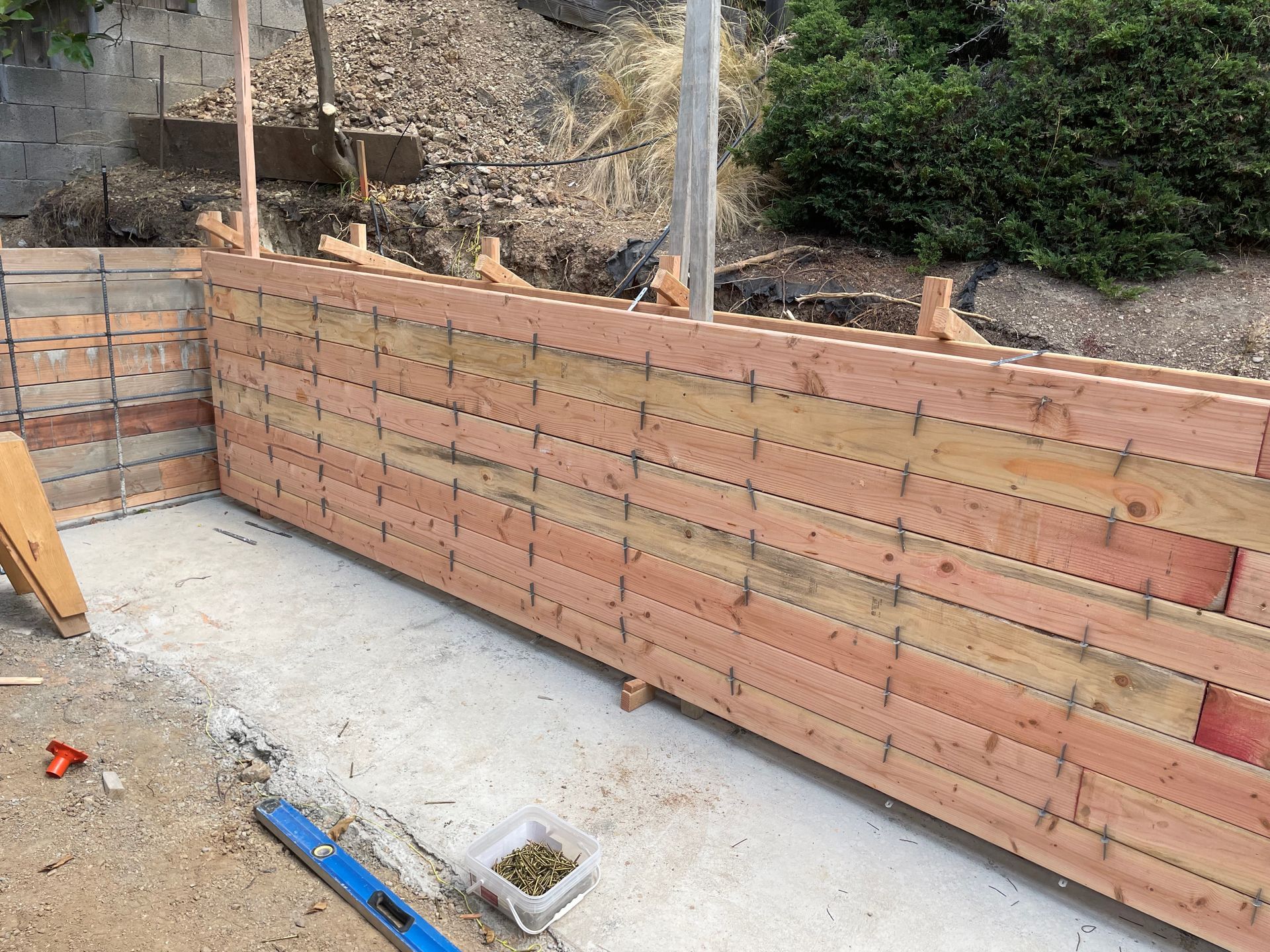 Wooden retaining wall under construction on a concrete foundation beside a dirt slope.