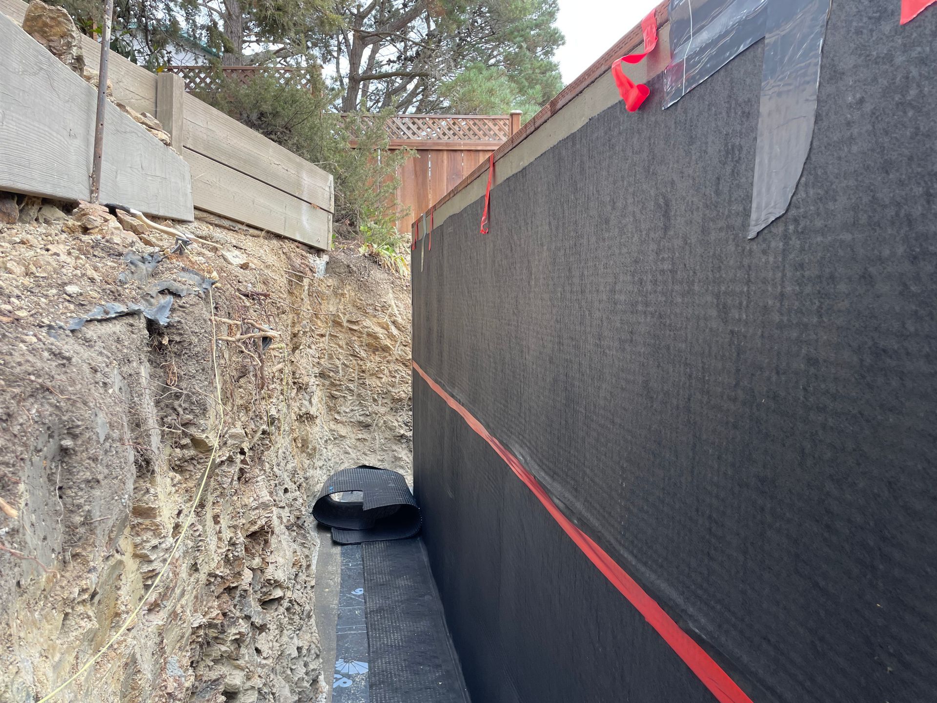 Narrow construction trench with rocky wall, black mesh fencing, and a drainage pipe along the bottom.
