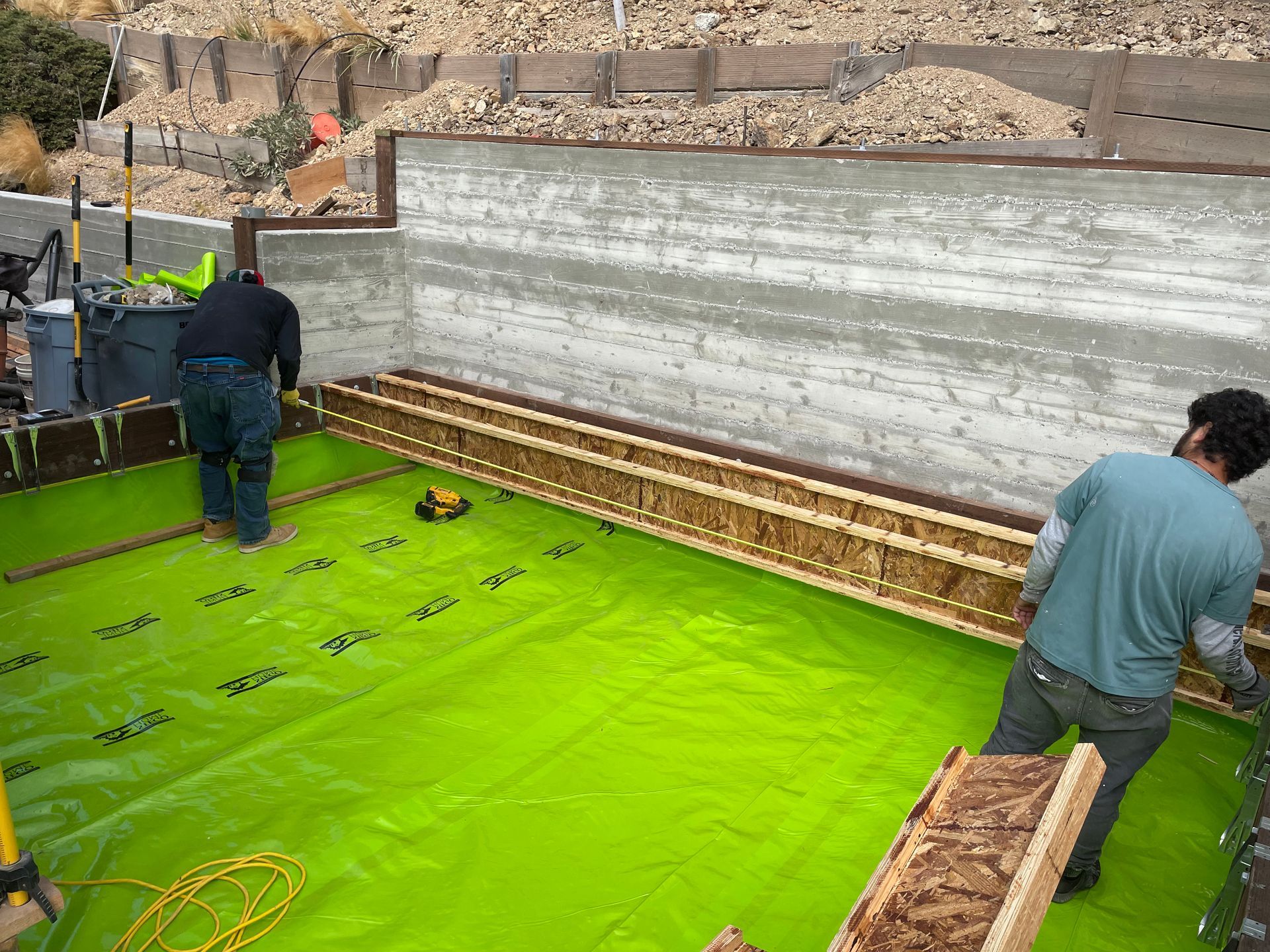 Workers installing bright green waterproofing on a construction foundation area.