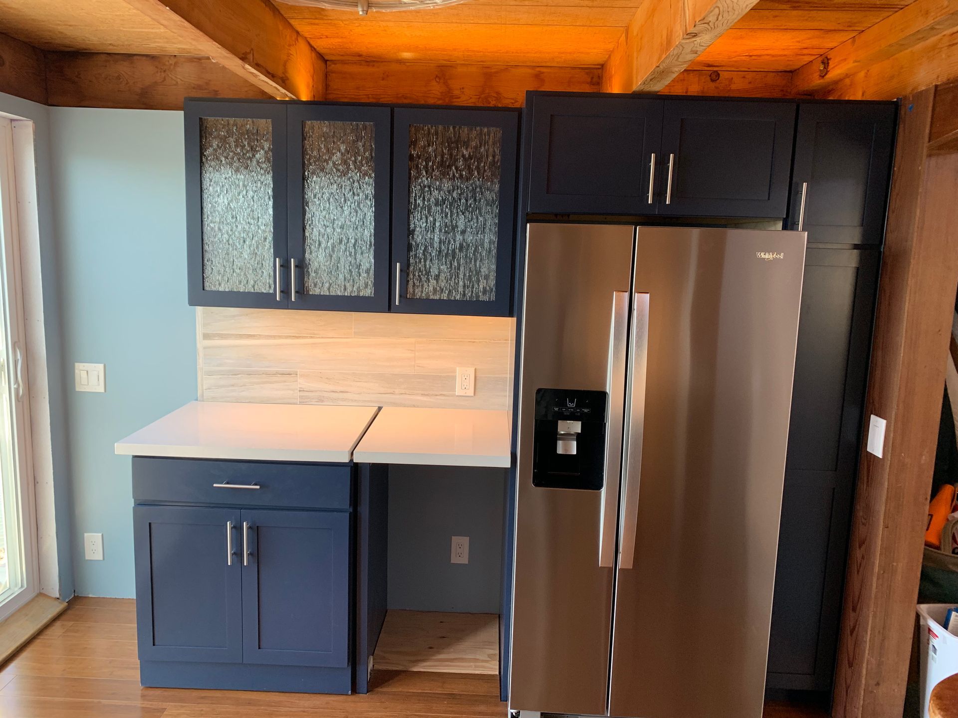 Modern kitchen corner with blue cabinets, white countertops, and stainless steel refrigerator under wood ceiling