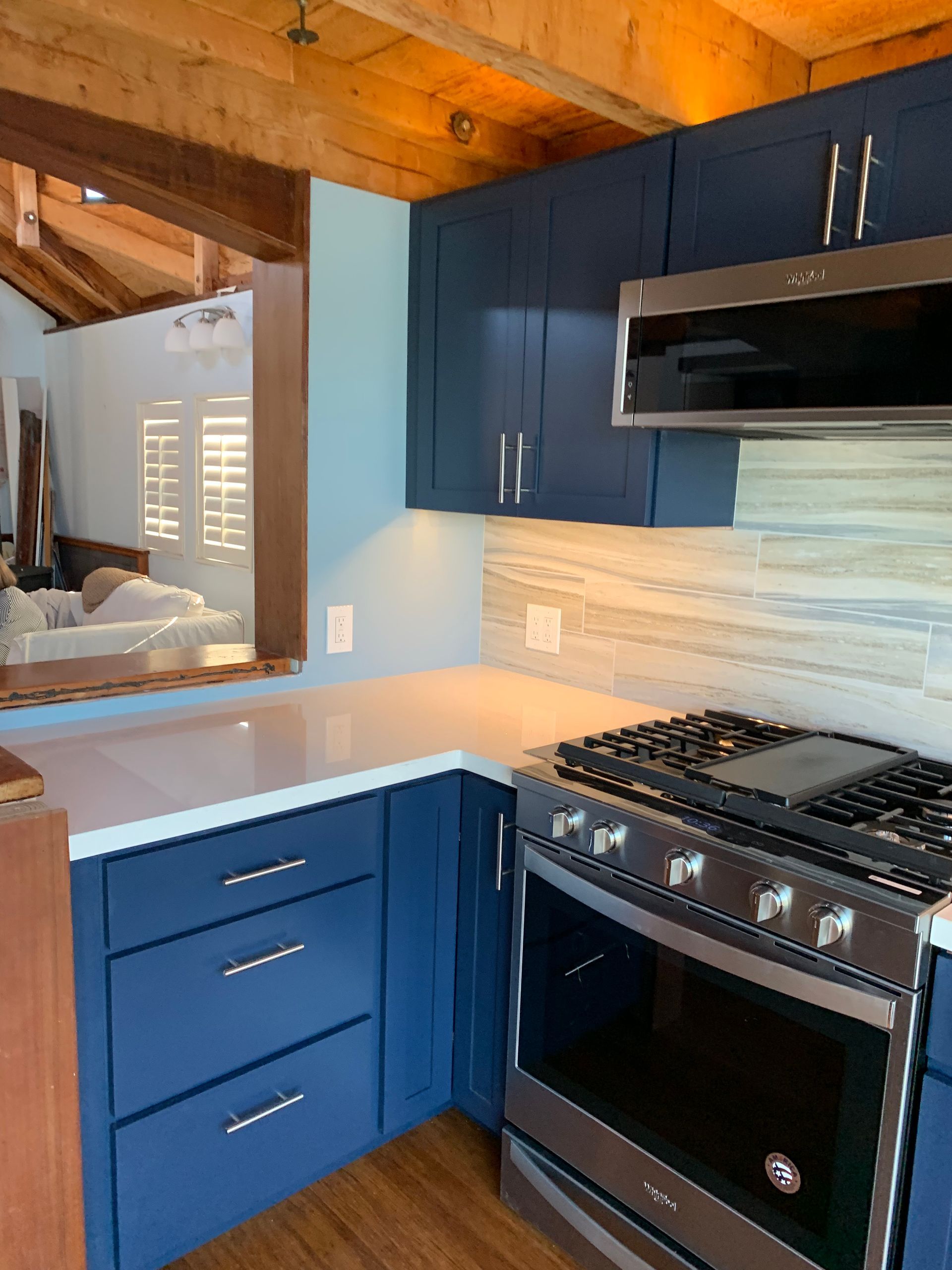 Blue kitchen with white countertops, stainless gas range, and wood ceiling beams, viewed through a pass-through opening