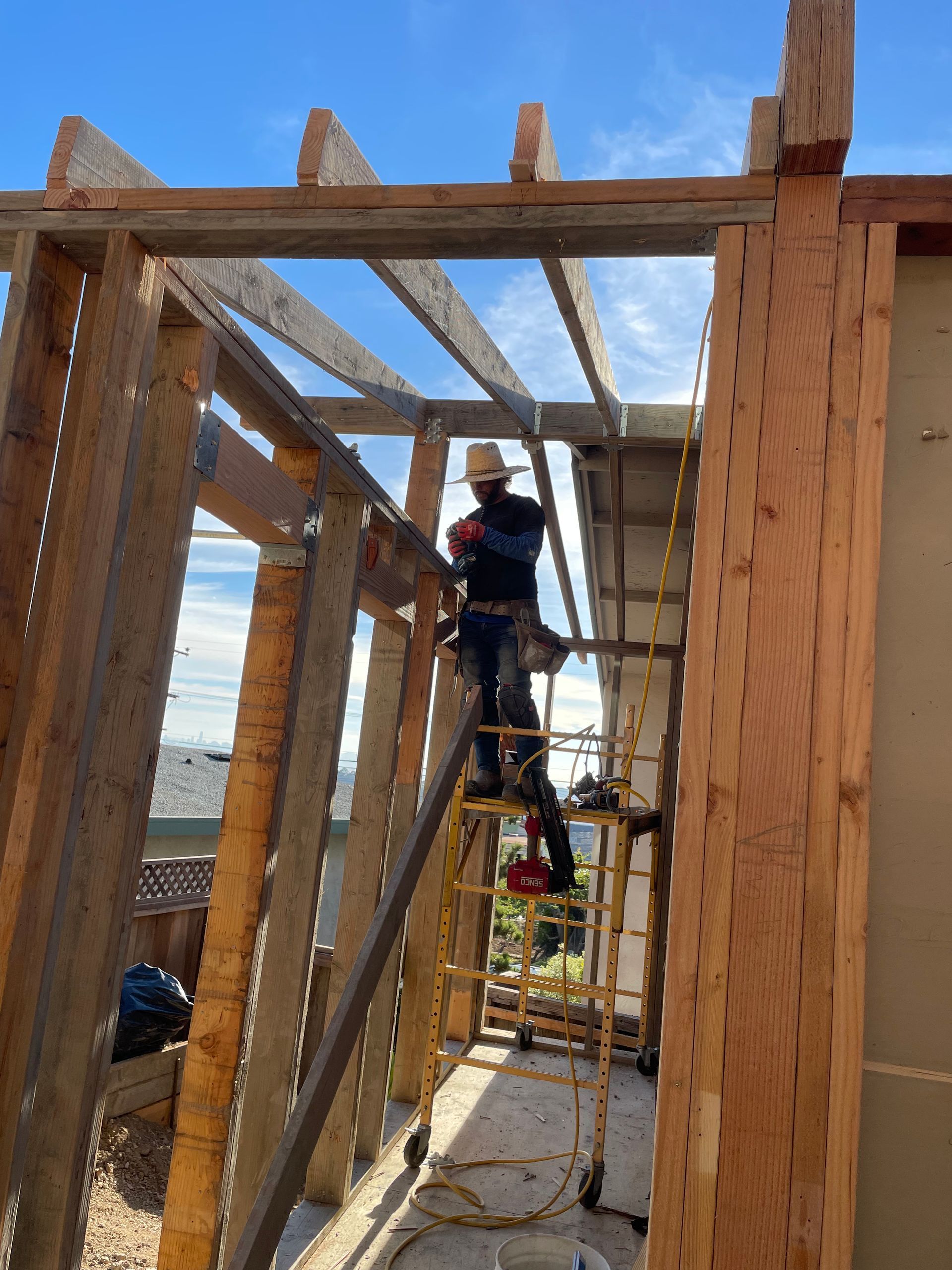 Worker on scaffolding inside a wooden building frame, wearing a harness and using a power tool.