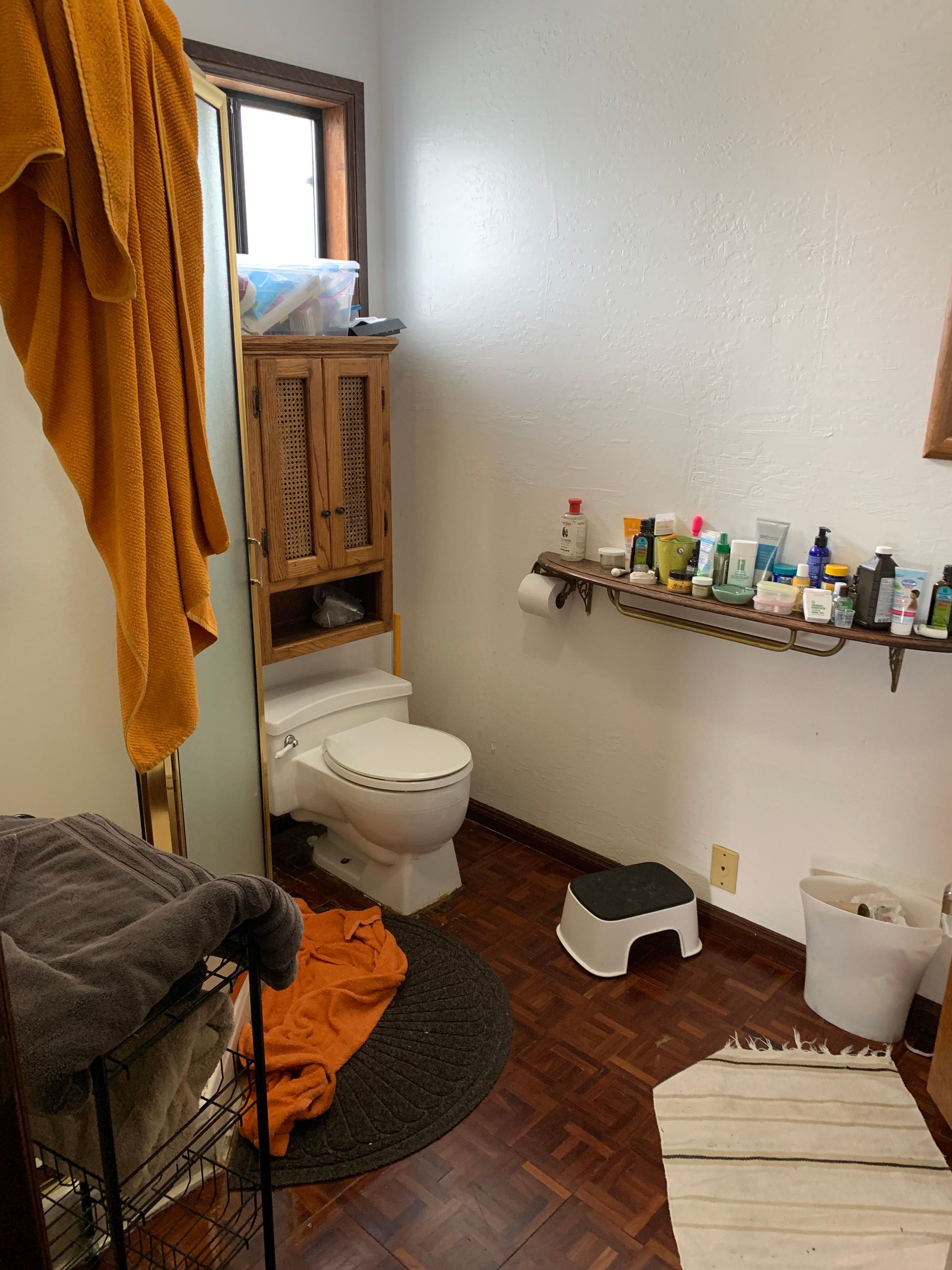 Small bathroom with toilet, towel rack, toiletries shelf, hamper, step stool, and striped rug.
