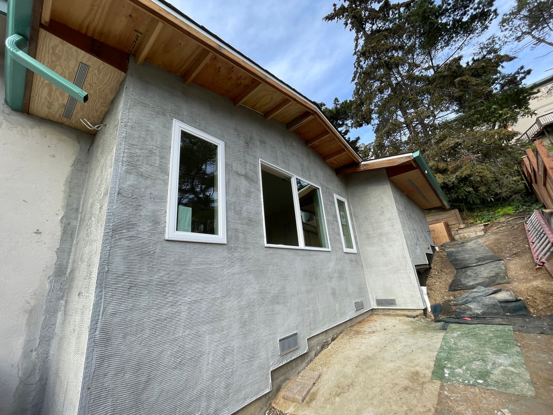 Gray stucco house exterior with white-framed windows, wooden eaves, and a narrow concrete side yard by stairs