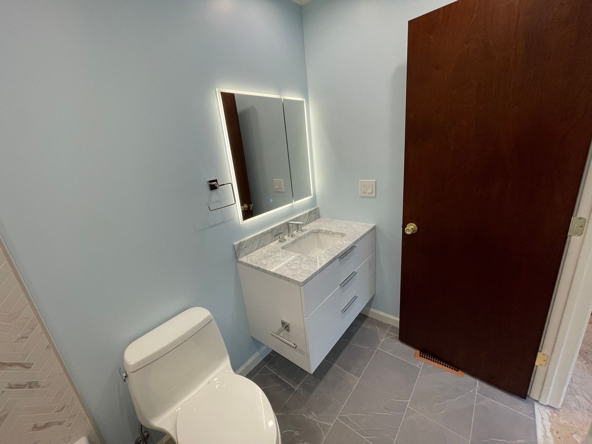 Small bathroom with white toilet, vanity, mirror, gray tile floor, and an open wooden door.