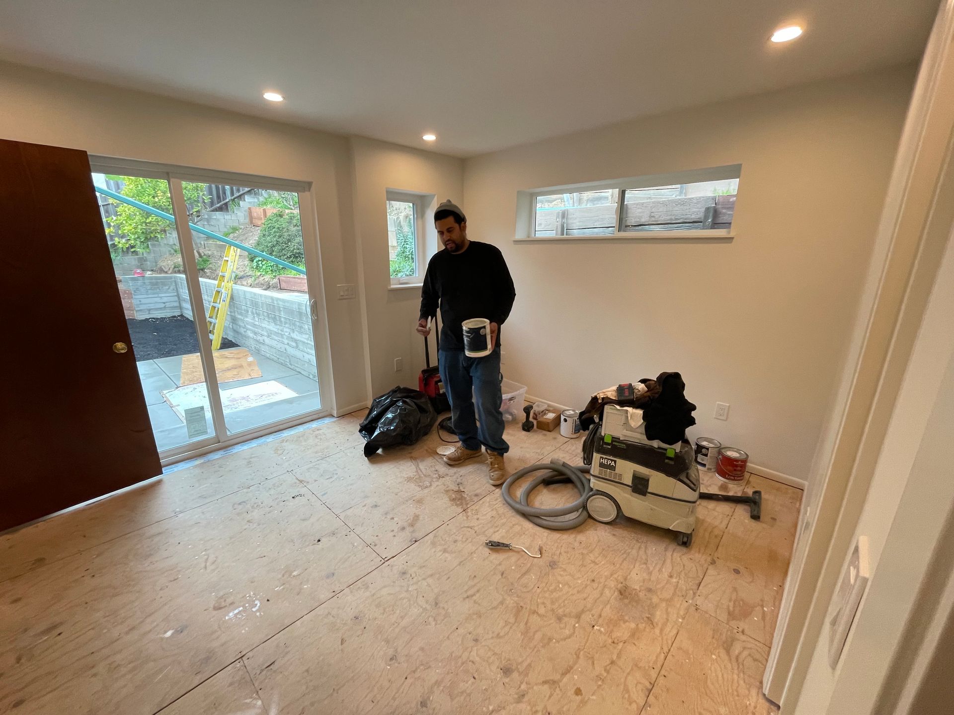 Person standing in a nearly empty room with tools and materials on the floor, near a large window and open door.