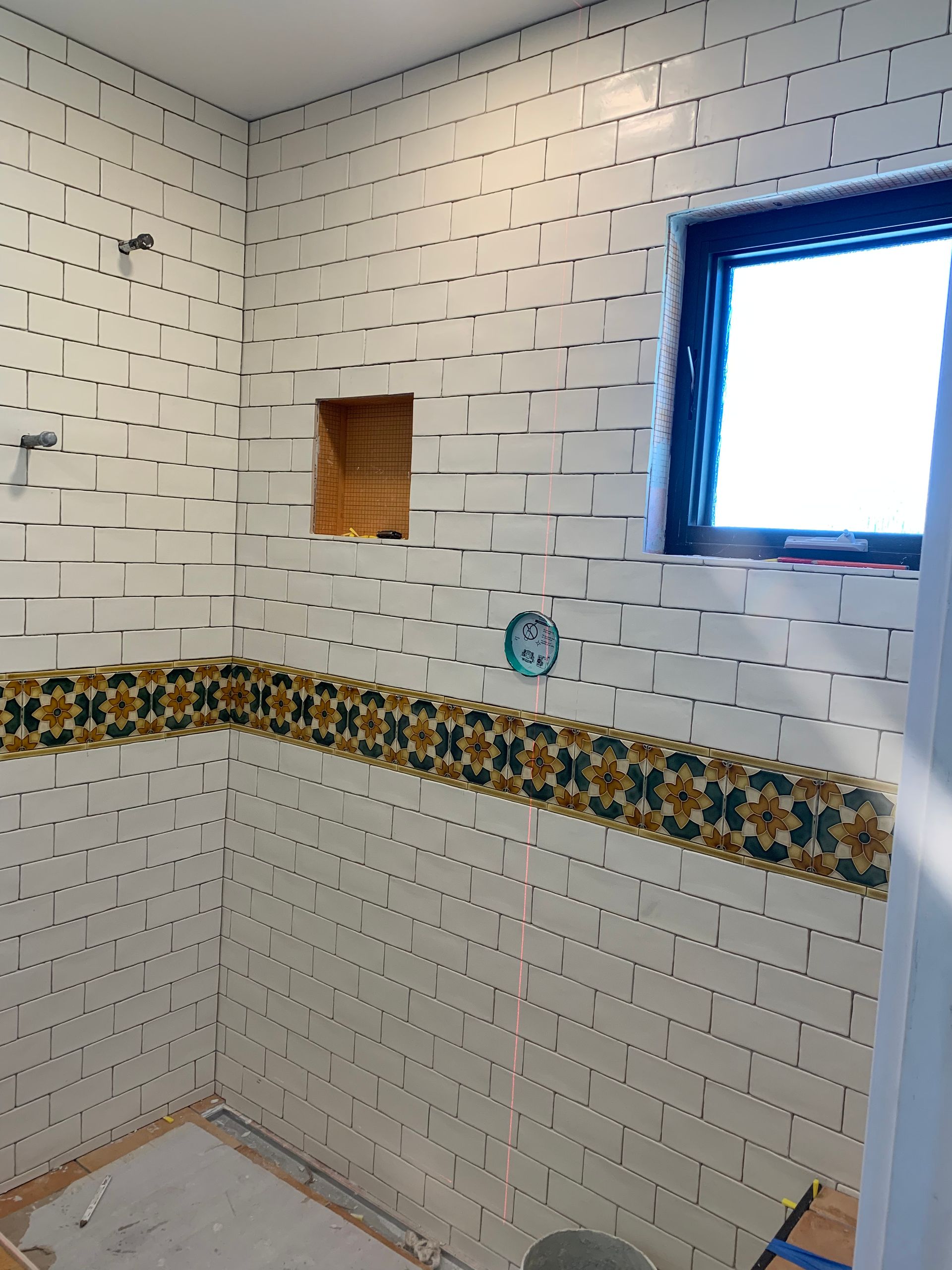 Tiled bathroom shower under construction with decorative border, recessed niche, and blue-framed window
