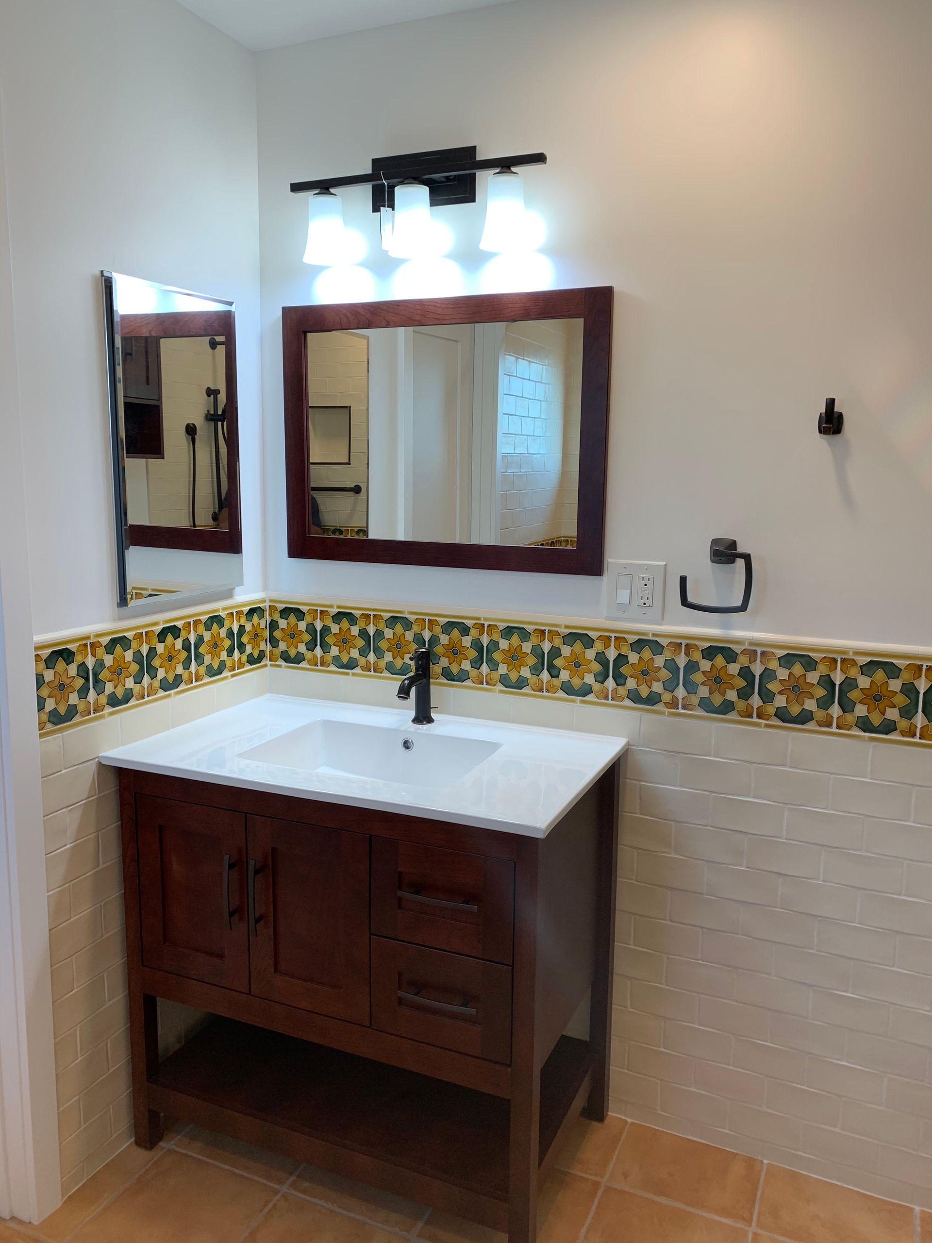 Small bathroom vanity with wood cabinet, white sink, mirror, and floral tile backsplash under a wall light.
