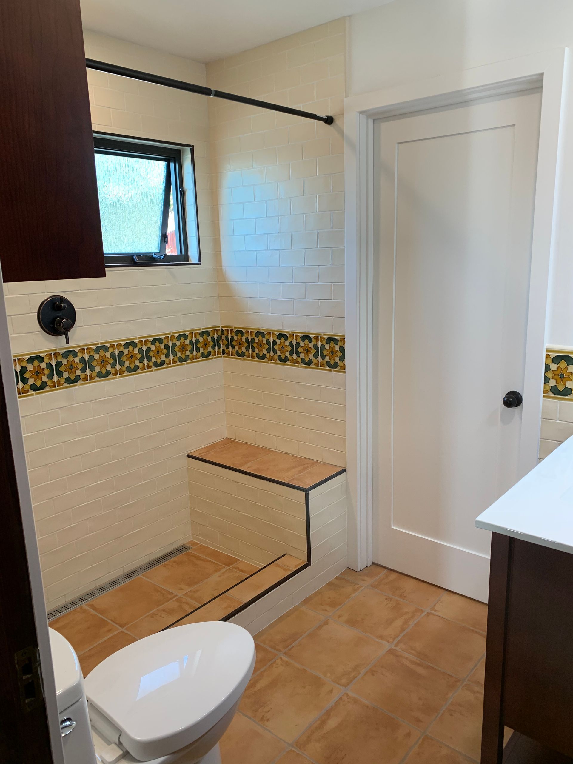 Small bathroom with a toilet, tiled shower bench, window, and white door.