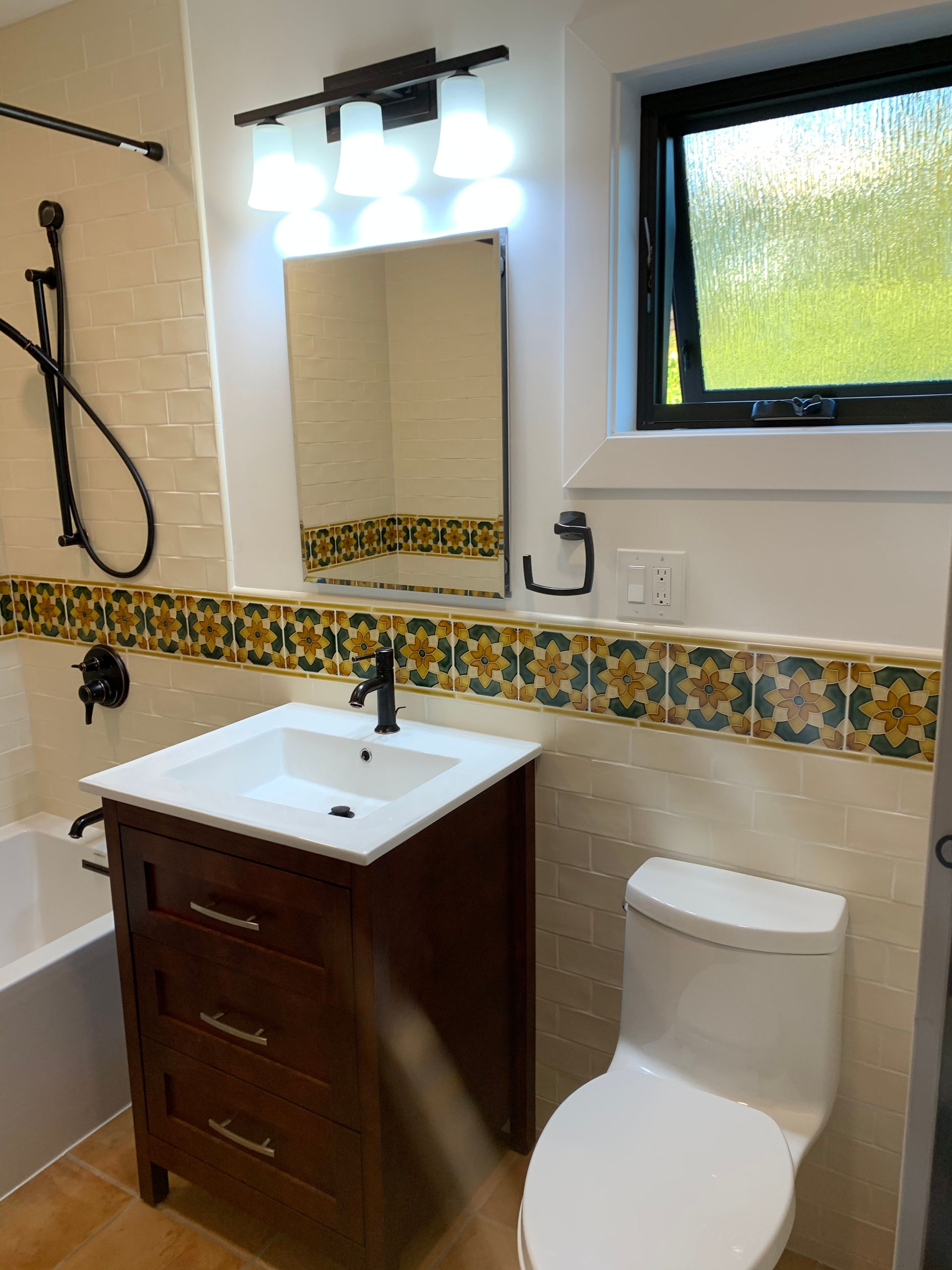 Small bathroom with white sink, wood vanity, toilet, mirror lights, and decorative tile trim.