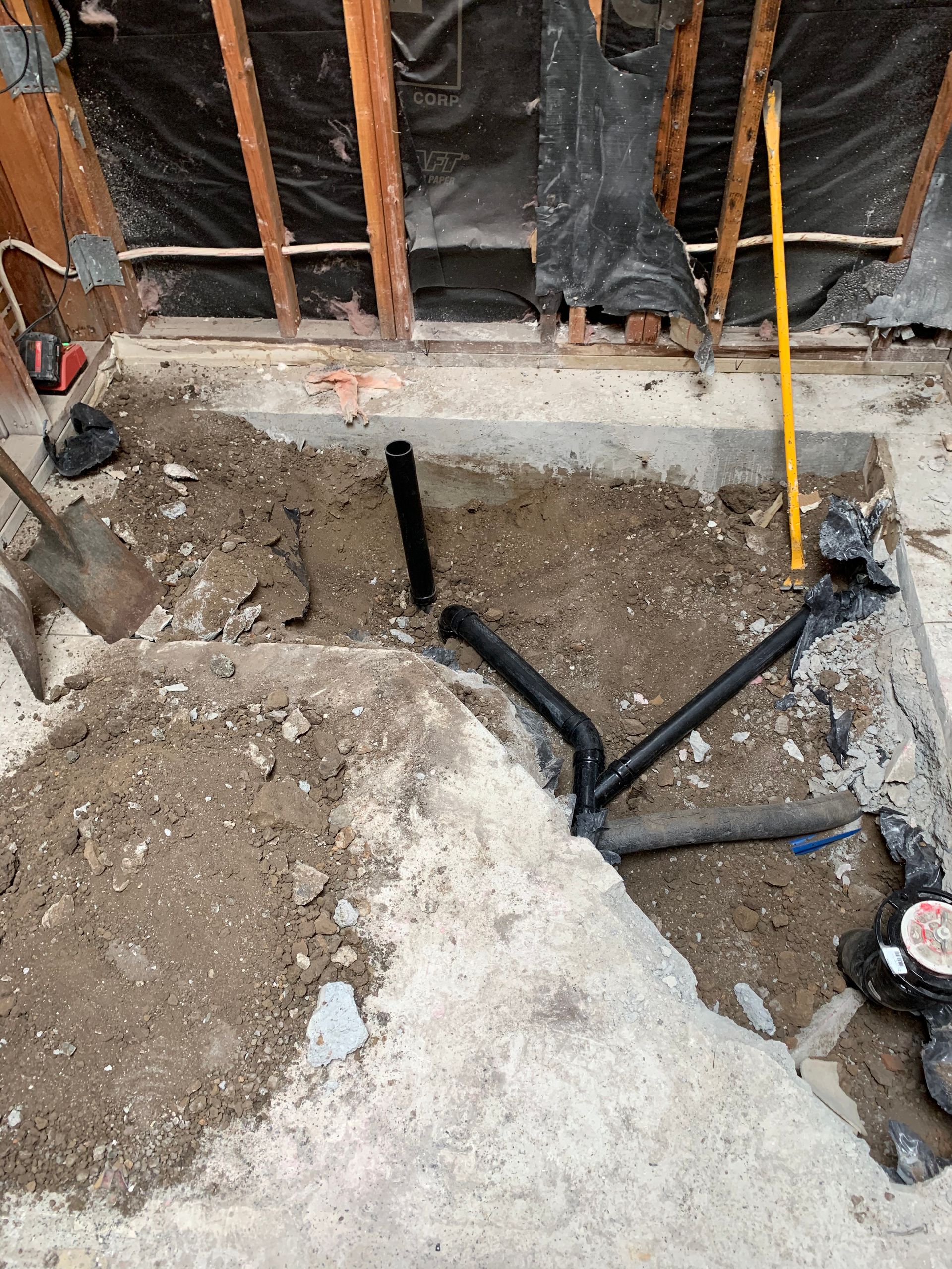 Construction hole in concrete floor with exposed pipes and debris inside a wall-framed room