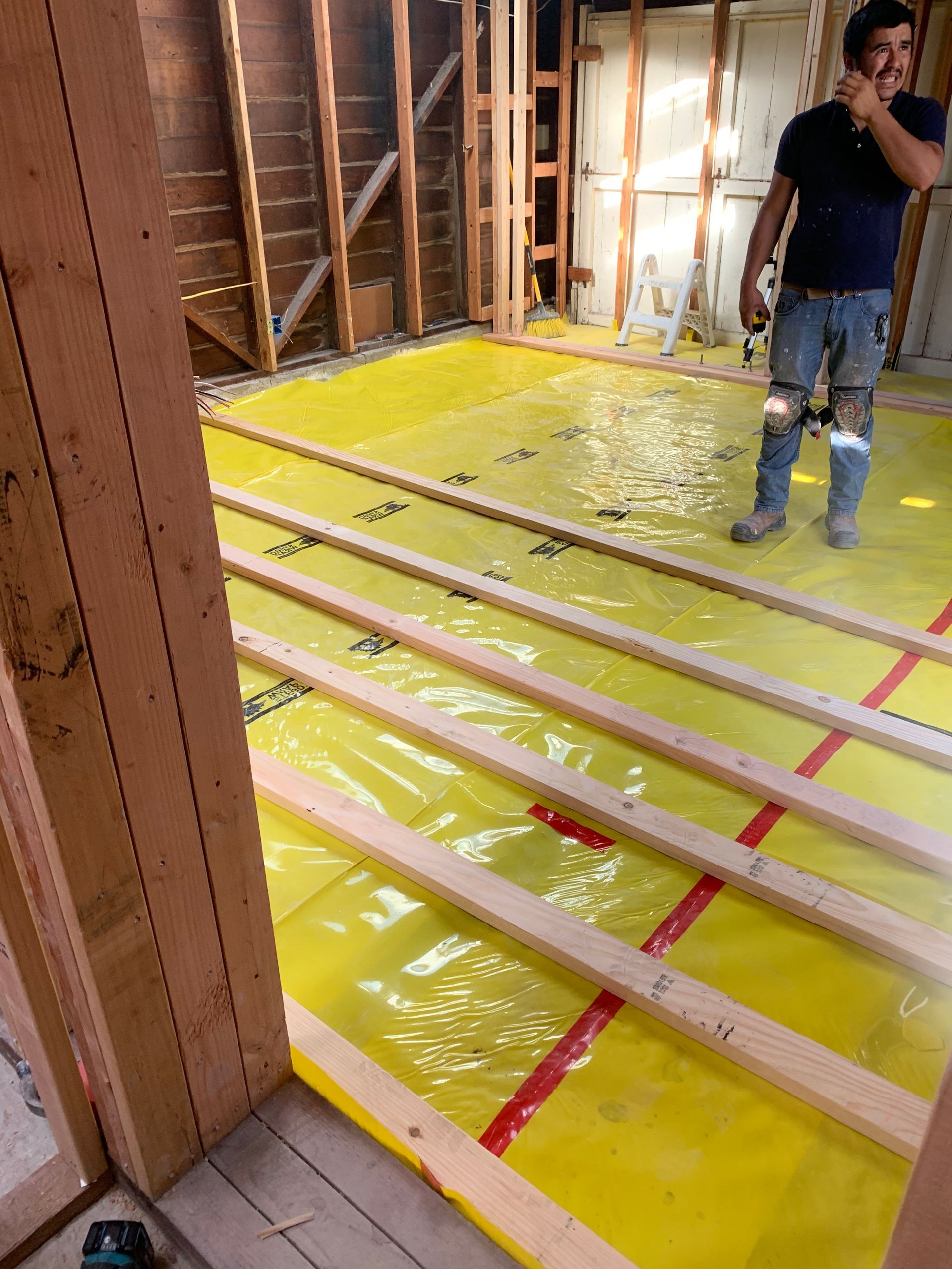 Unfinished room with yellow floor covering, exposed joists, and a worker standing near the wall.