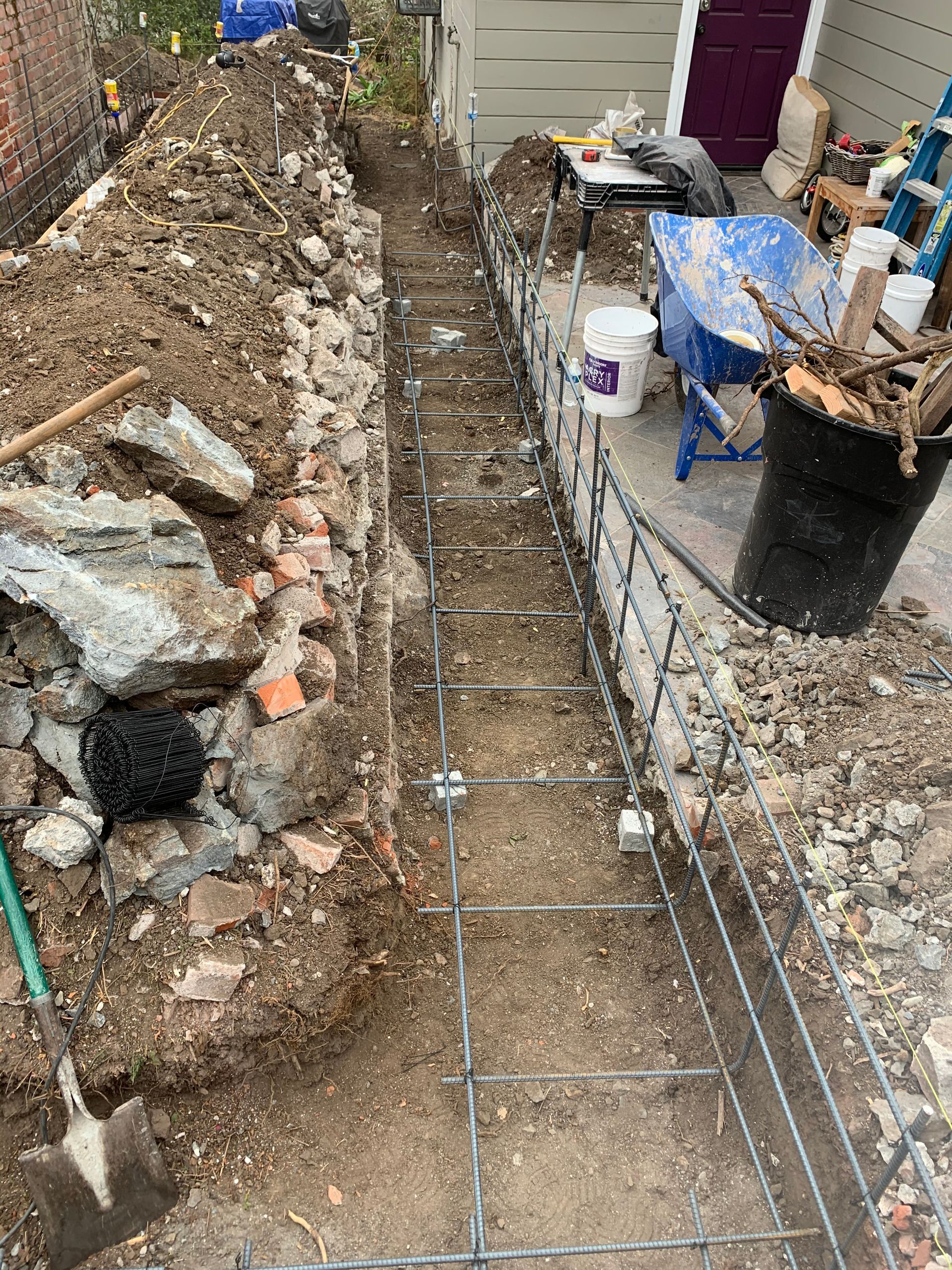 Narrow trench beside a house, with rebar, rubble, and a work bucket on the right side.
