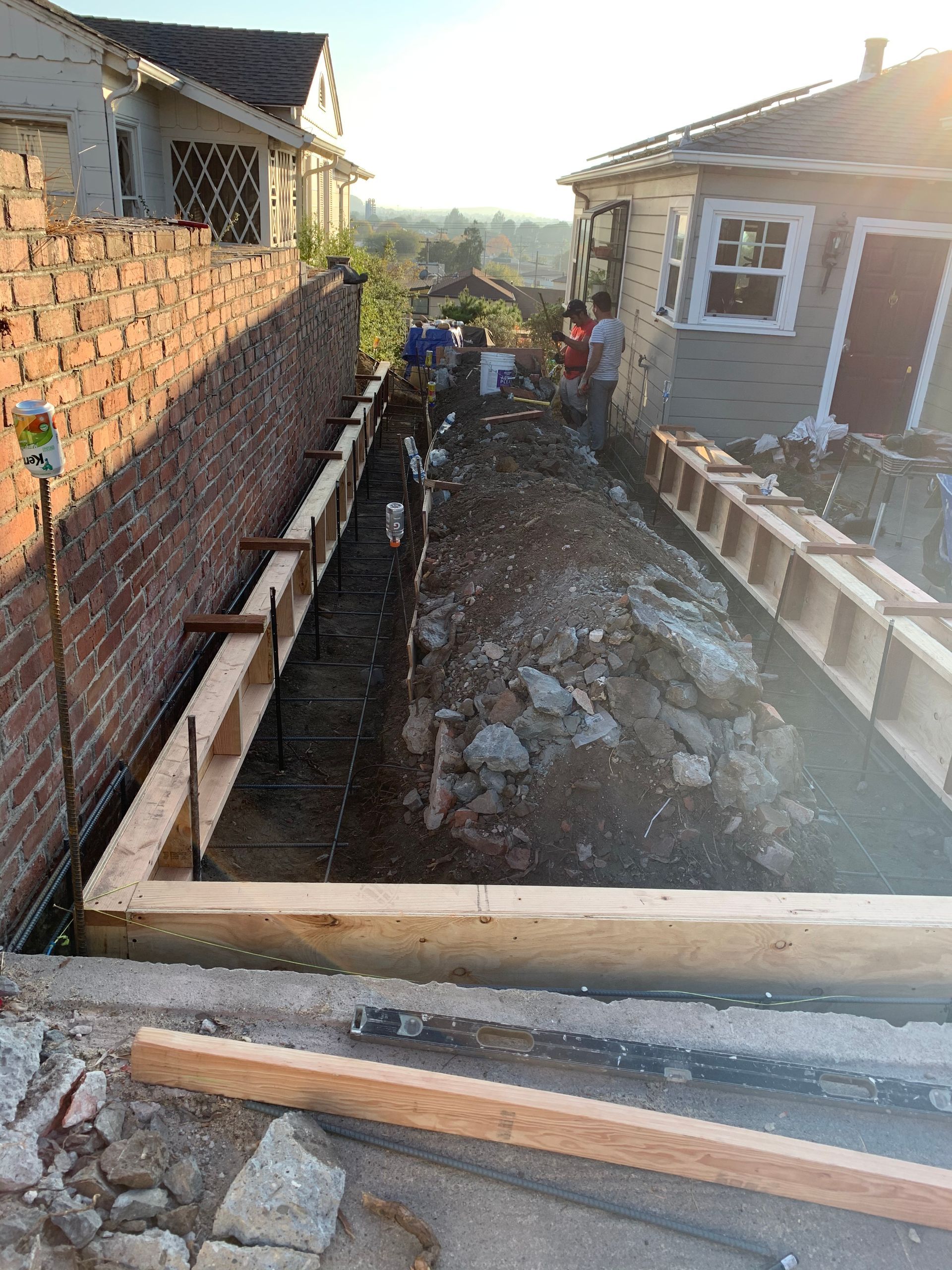 Backyard construction site with wooden framing, rubble, and a narrow trench between brick and house walls