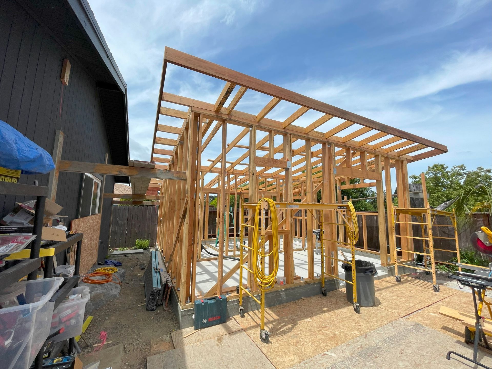 Wood-framed house under construction beside a dark building, with scaffolding and lumber on a sunny day