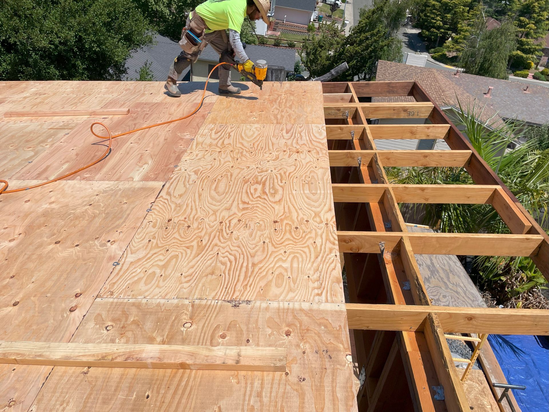 Worker installing plywood roof decking on a house frame with exposed rafters.