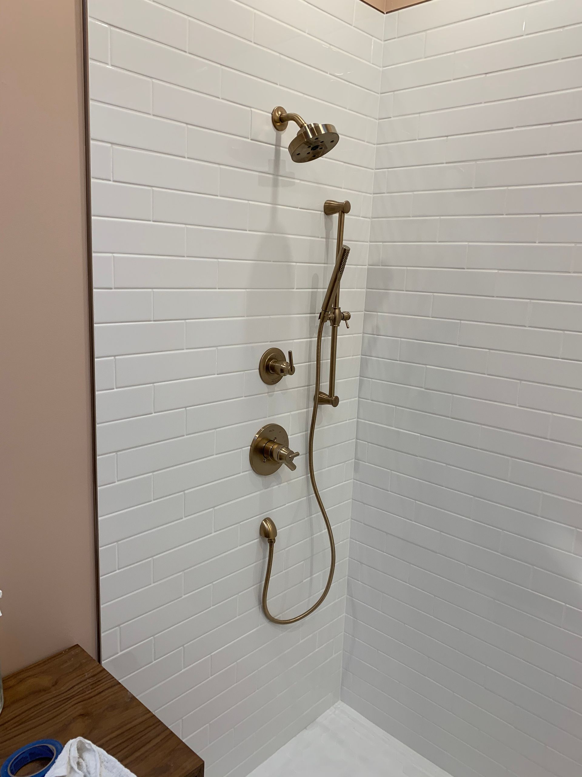 White-tiled walk-in shower with brass fixtures, handheld sprayer, and wooden floor outside.
