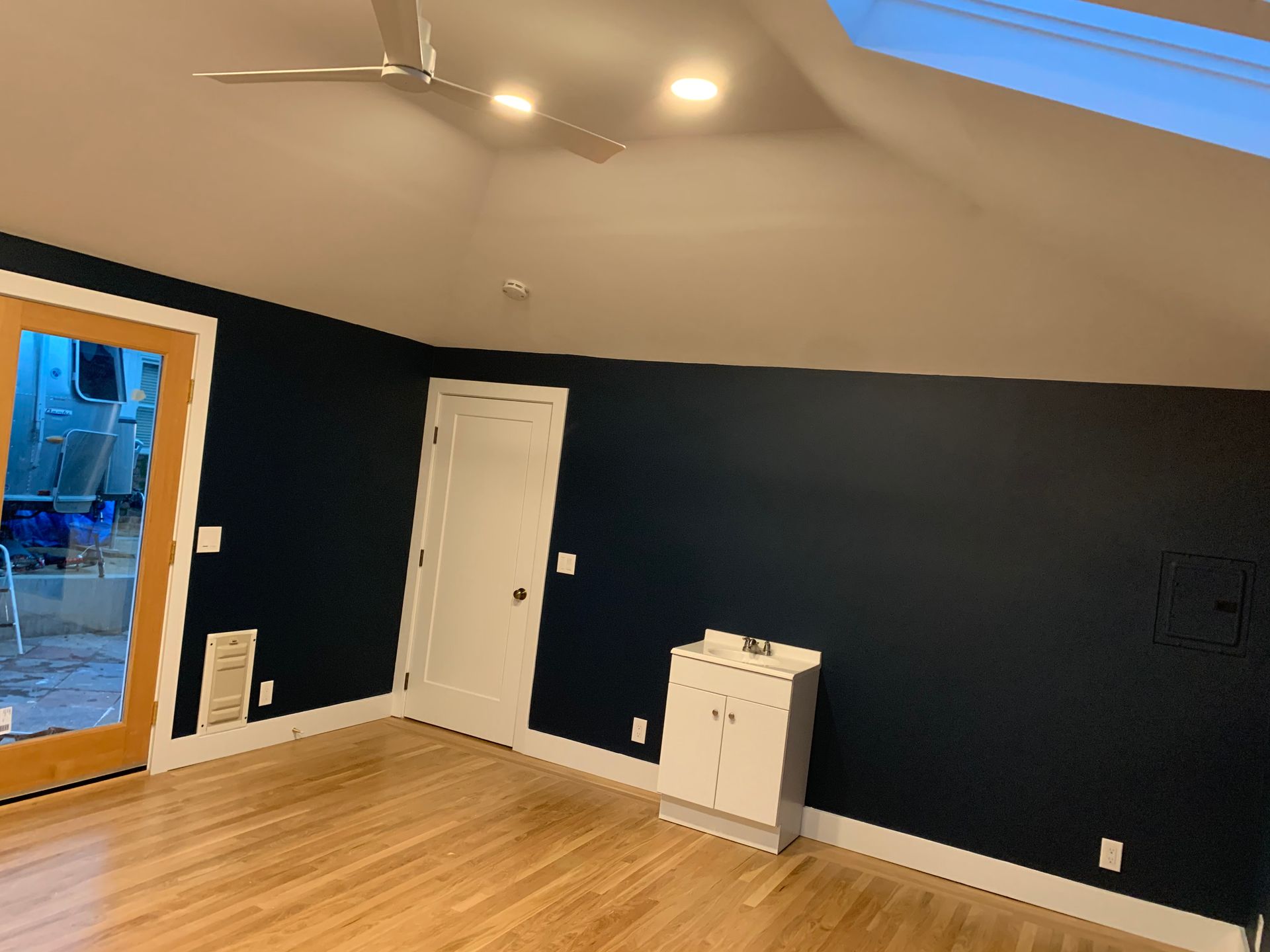 Empty room with dark blue walls, wood floor, white door, ceiling fan, and open doorway to outside.