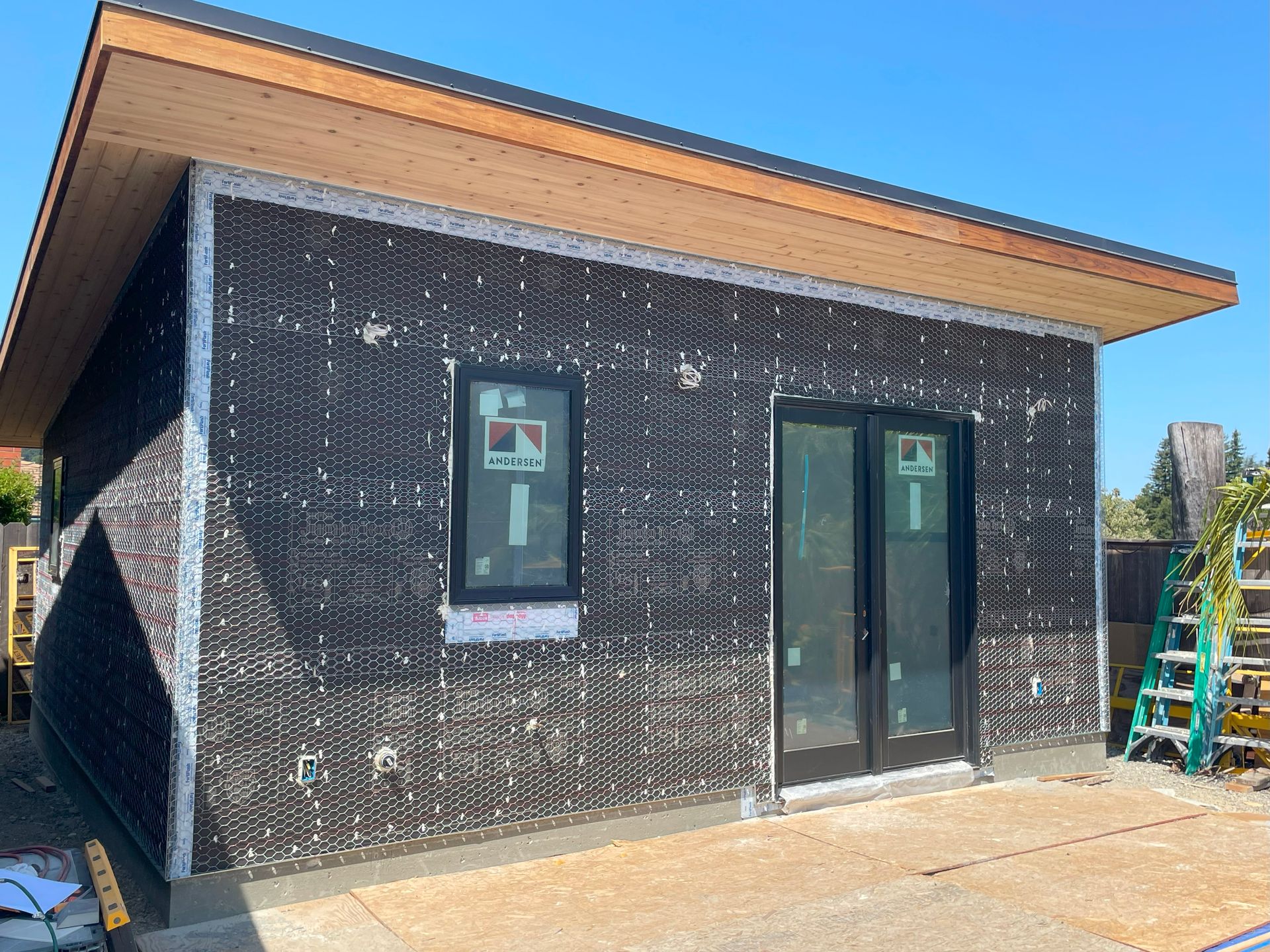 Small modern building exterior with black wrap, wood roof trim, two windows, and a glass door under construction
