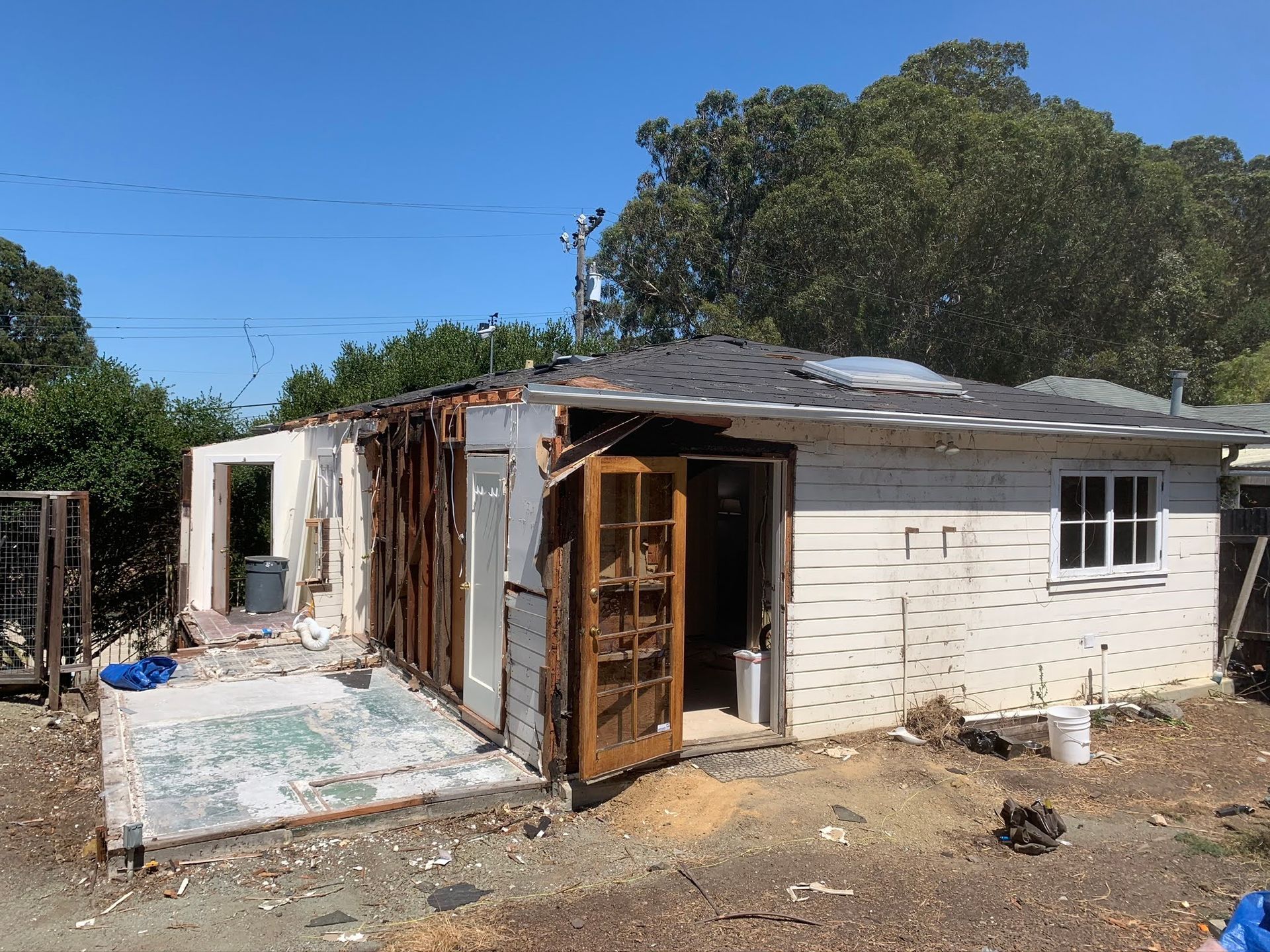 Damaged single-story house with broken doors and a patched roof in a dusty yard under a clear blue sky