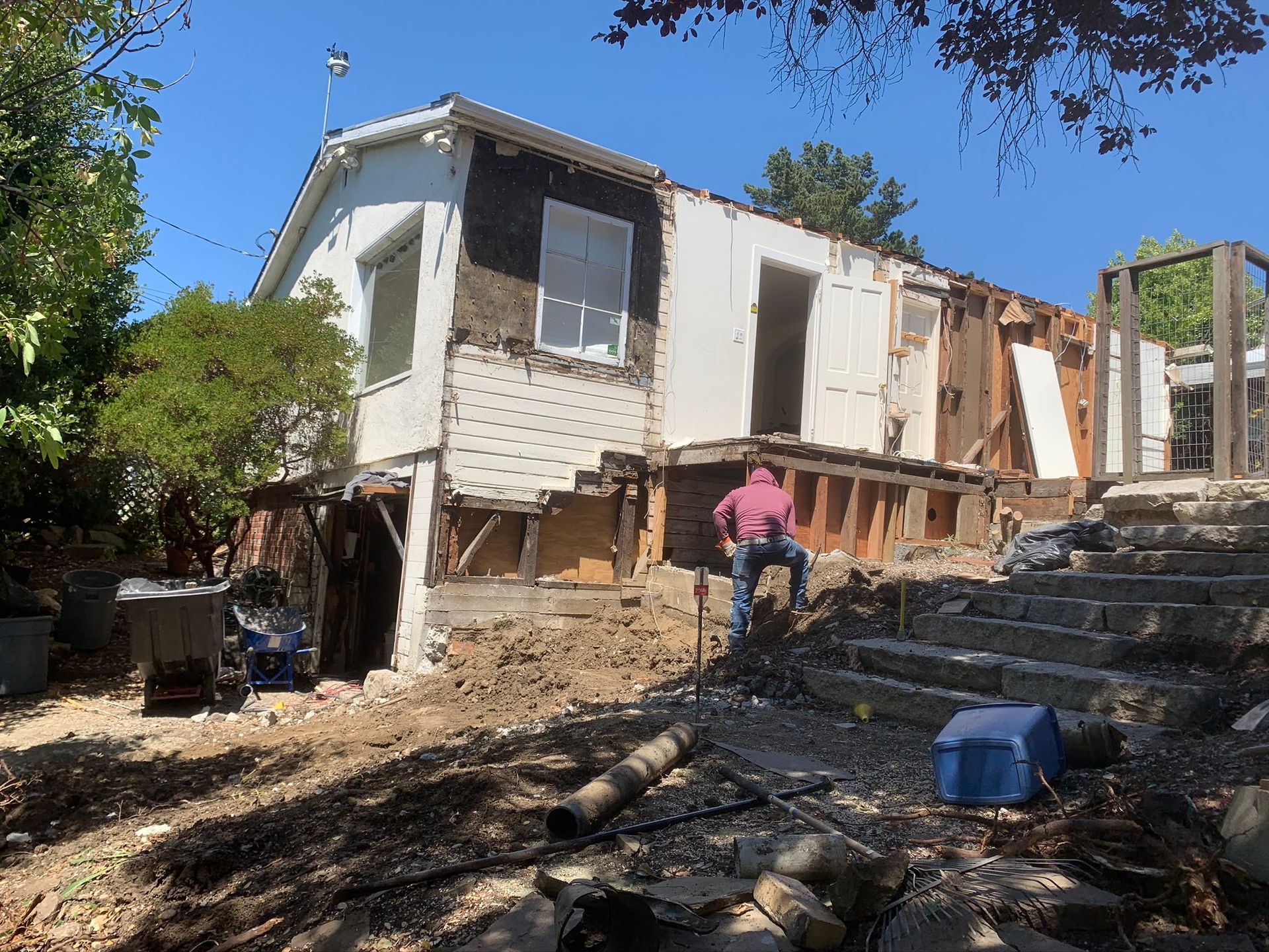 House under renovation with exposed framing, debris, and a worker on a sunny slope