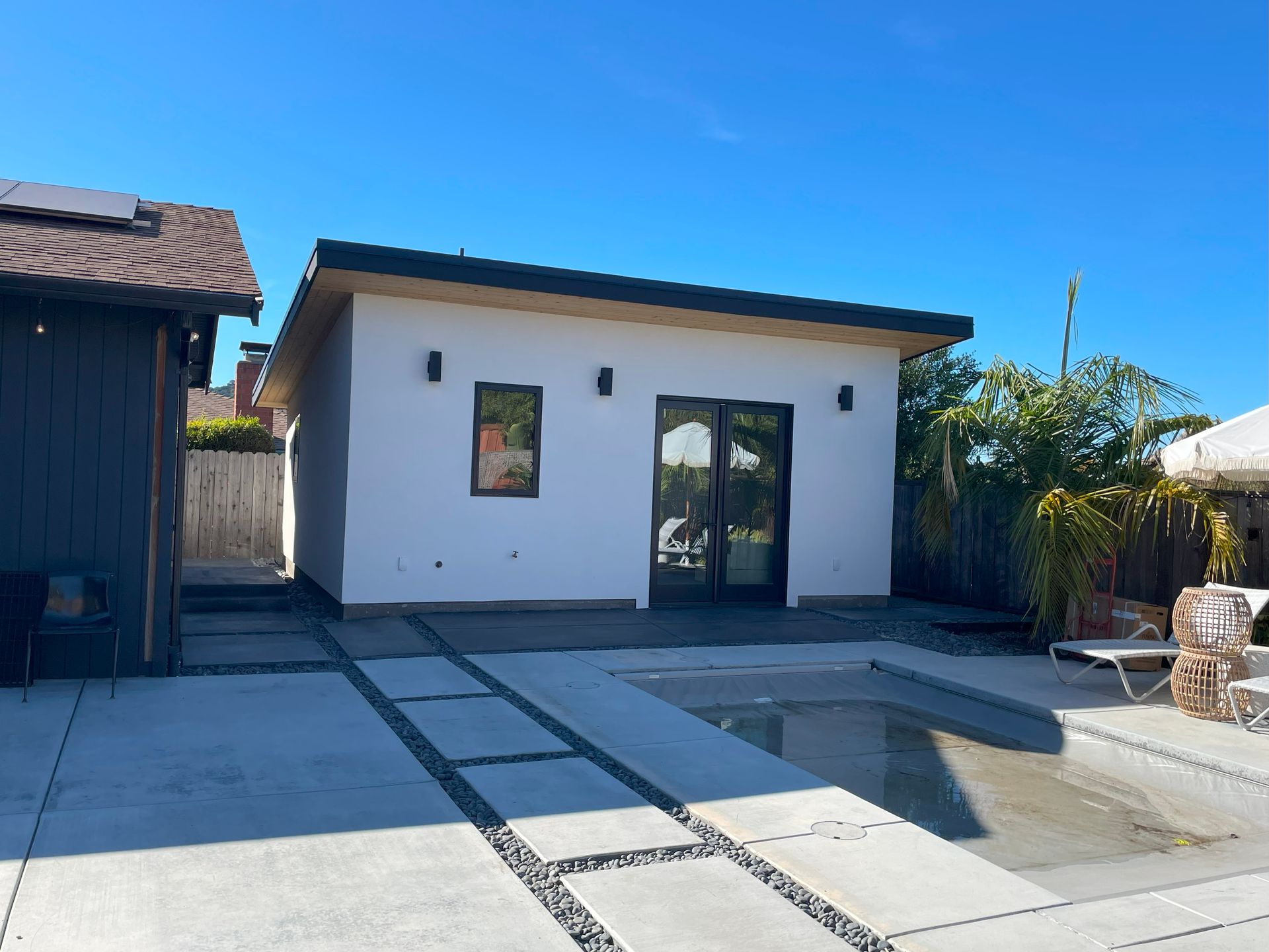 Backyard patio with a small white pool house, concrete deck, and a shallow pool area under a blue sky