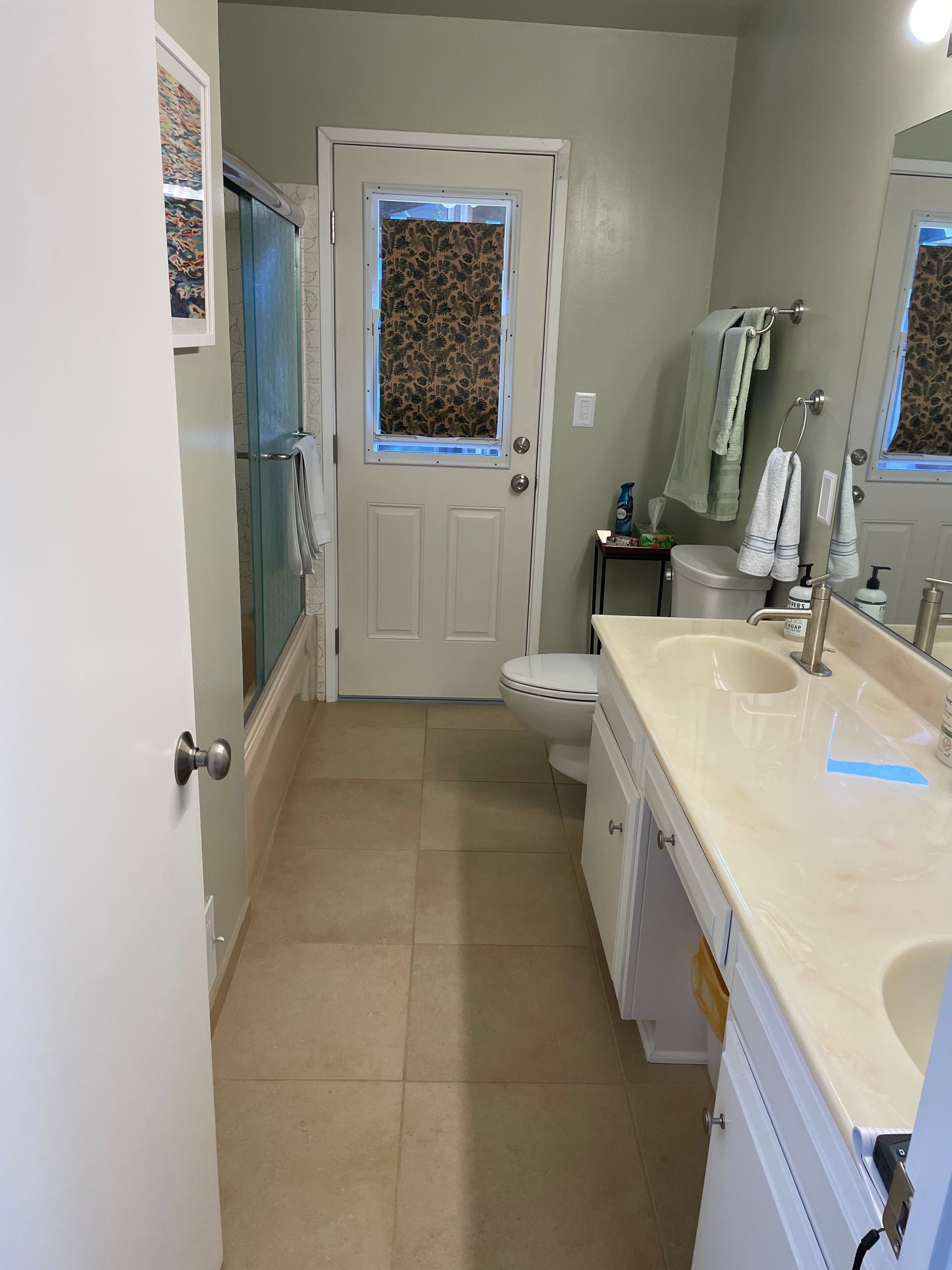 Narrow bathroom with beige tile, white vanity, toilet, and a door with a window at the end.