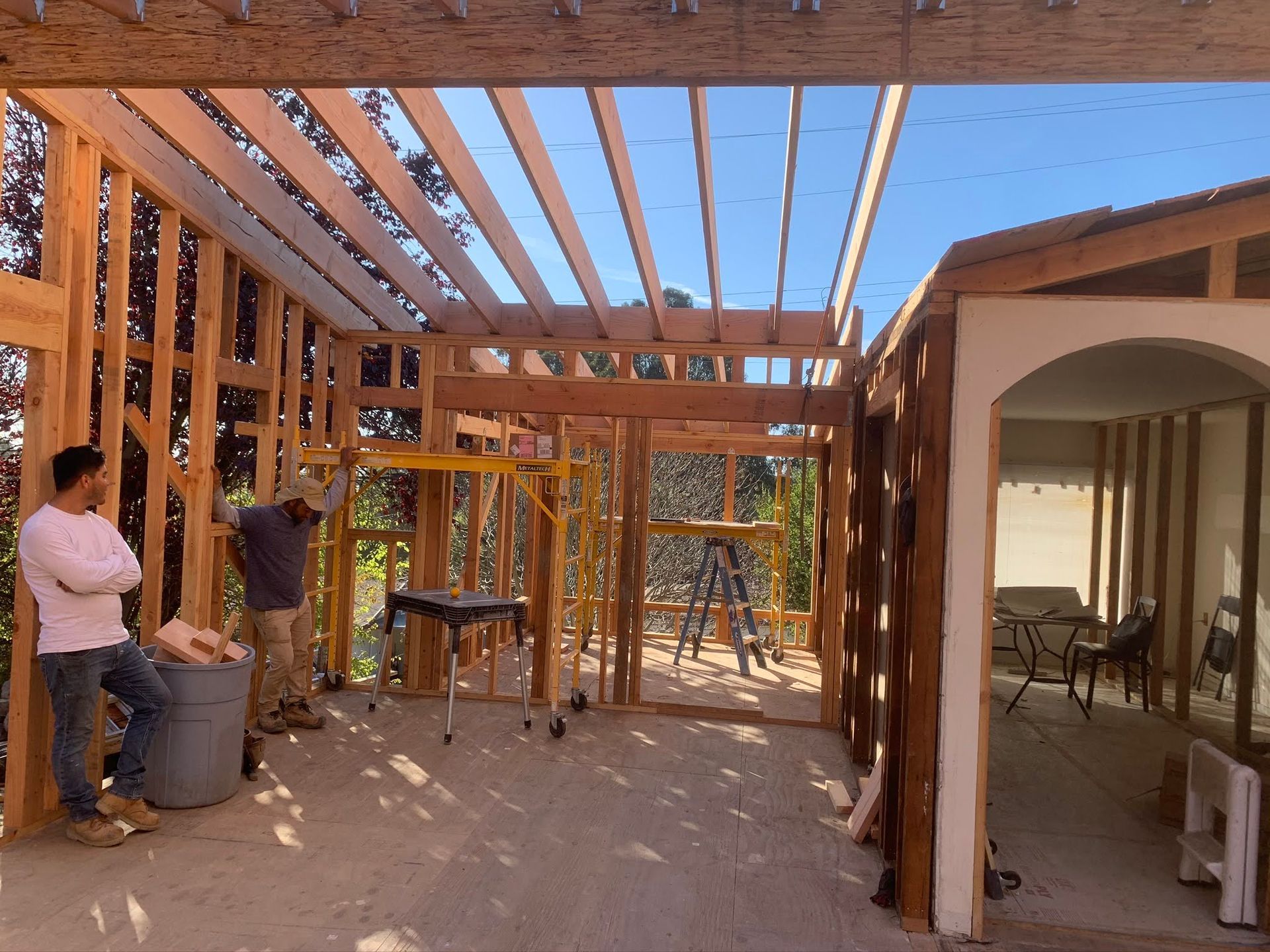 Wood-frame house under construction with workers and exposed beams in a sunlit yard