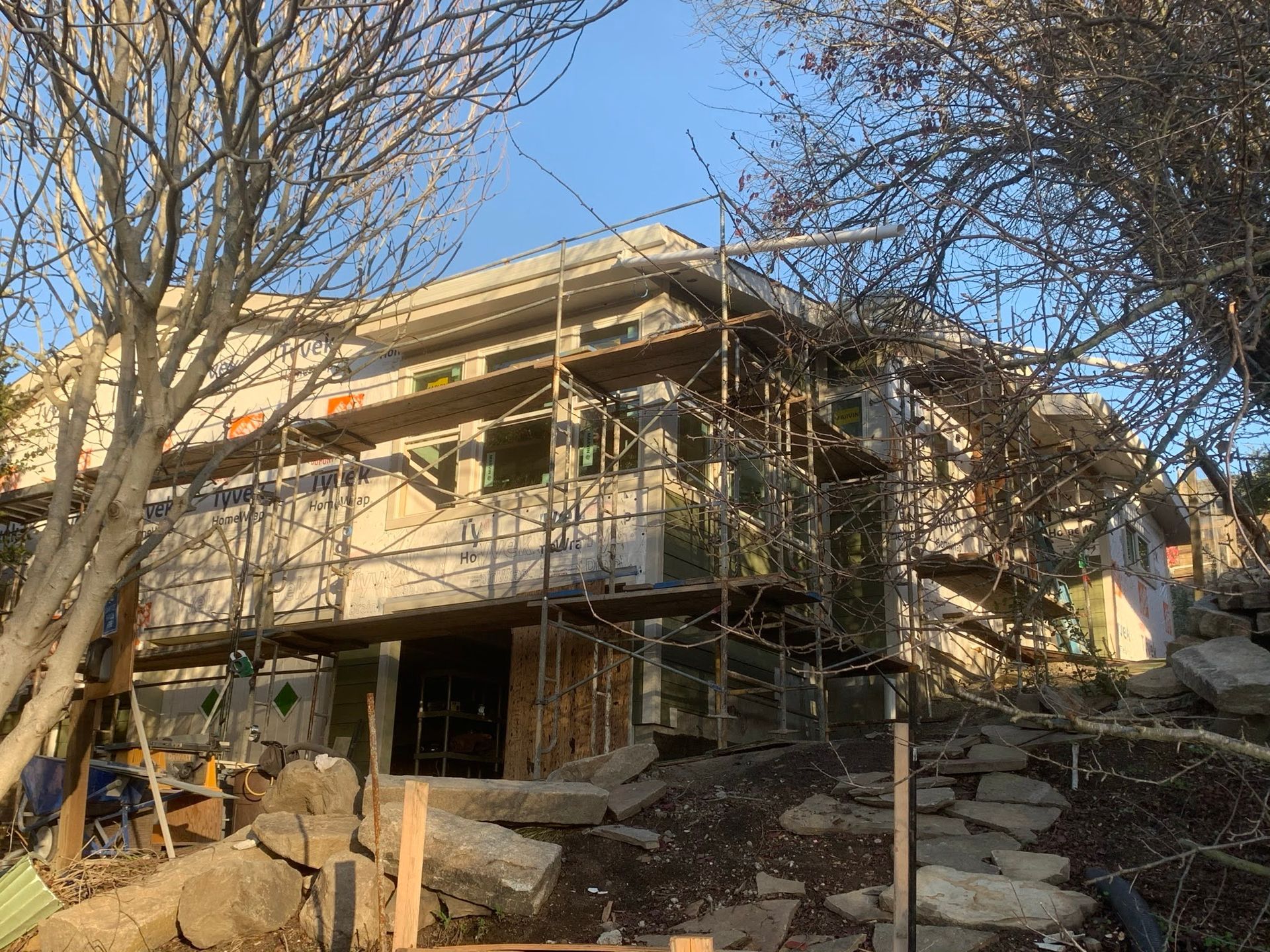 Two-story house under construction with scaffolding and exposed framing, surrounded by trees and rocks