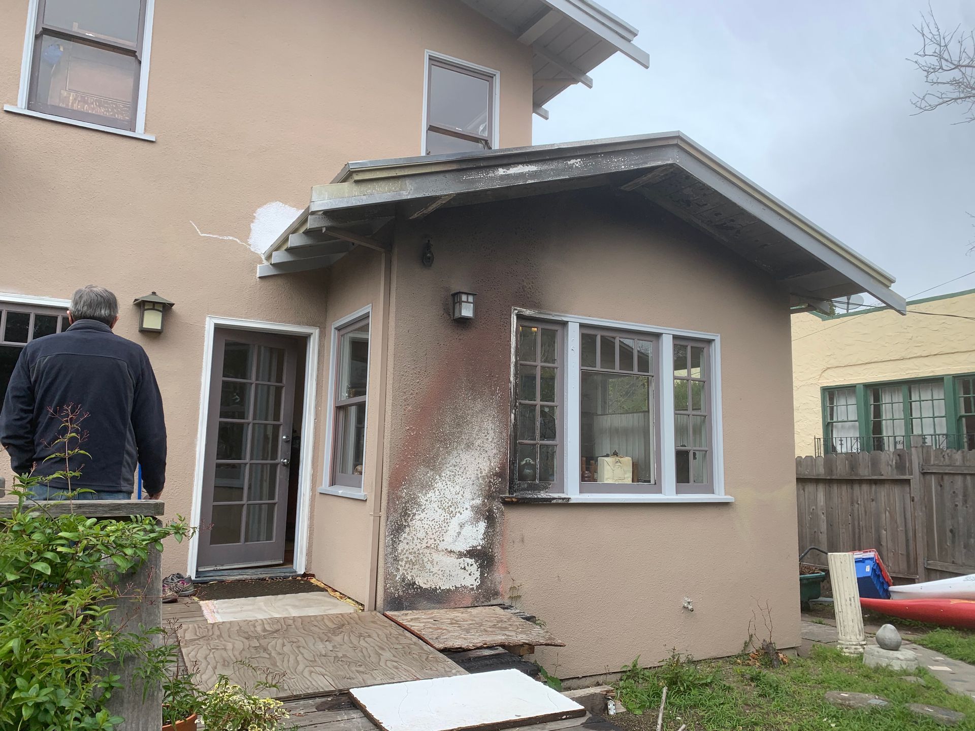Damaged beige house exterior with broken siding and a person standing by the back door