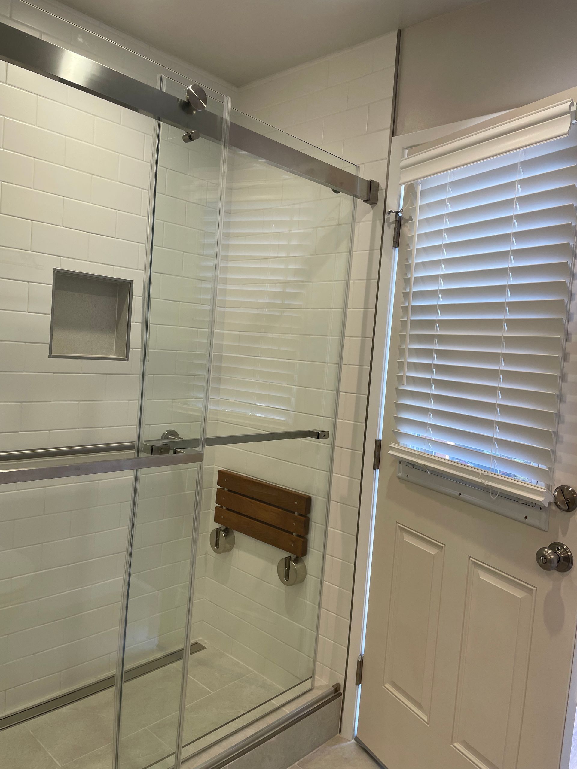 Glass shower enclosure beside a white bathroom door with blinds and a towel bar under the window