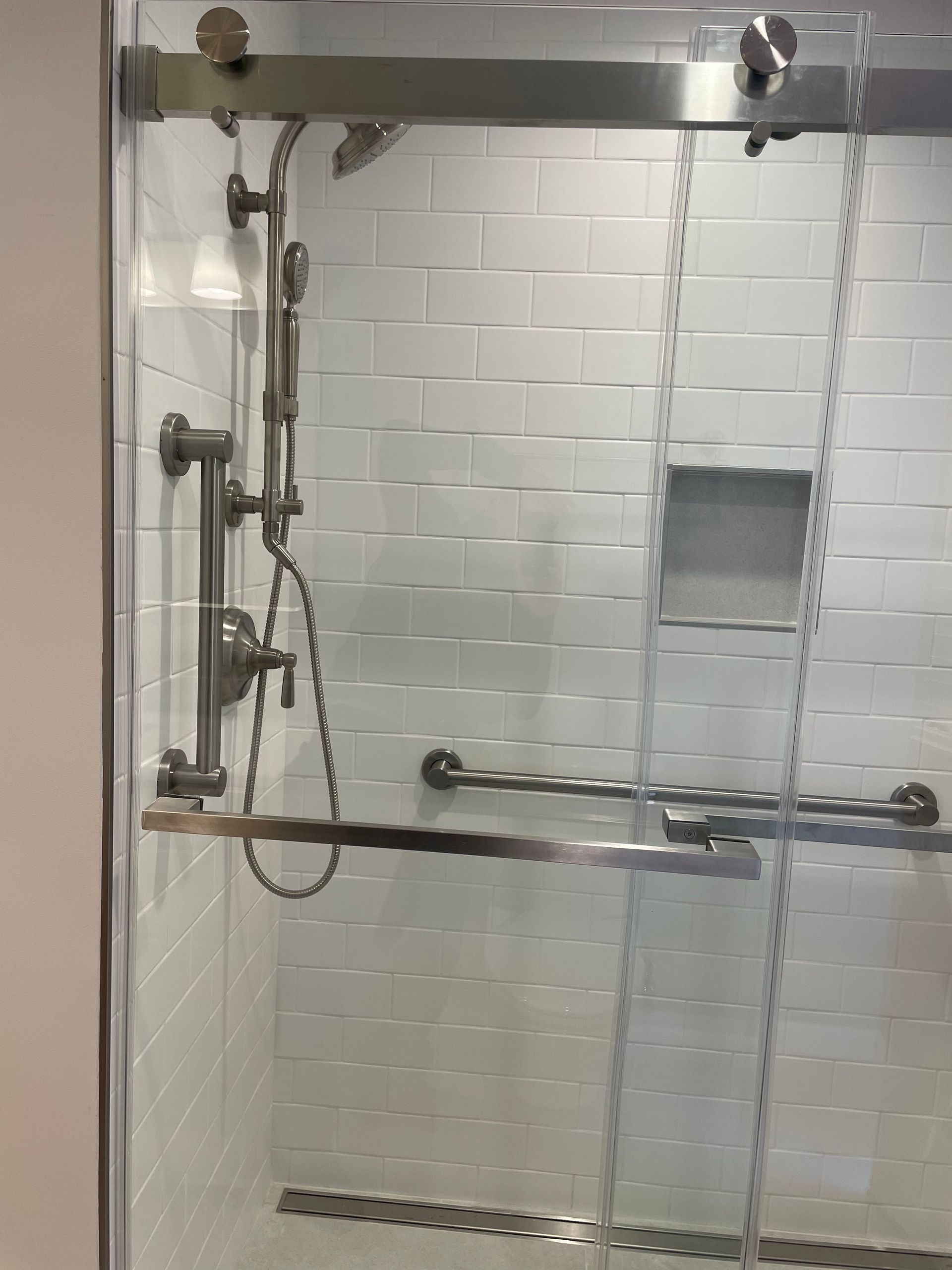 Accessible shower stall with grab bars, handheld shower, and built-in shelf behind glass doors