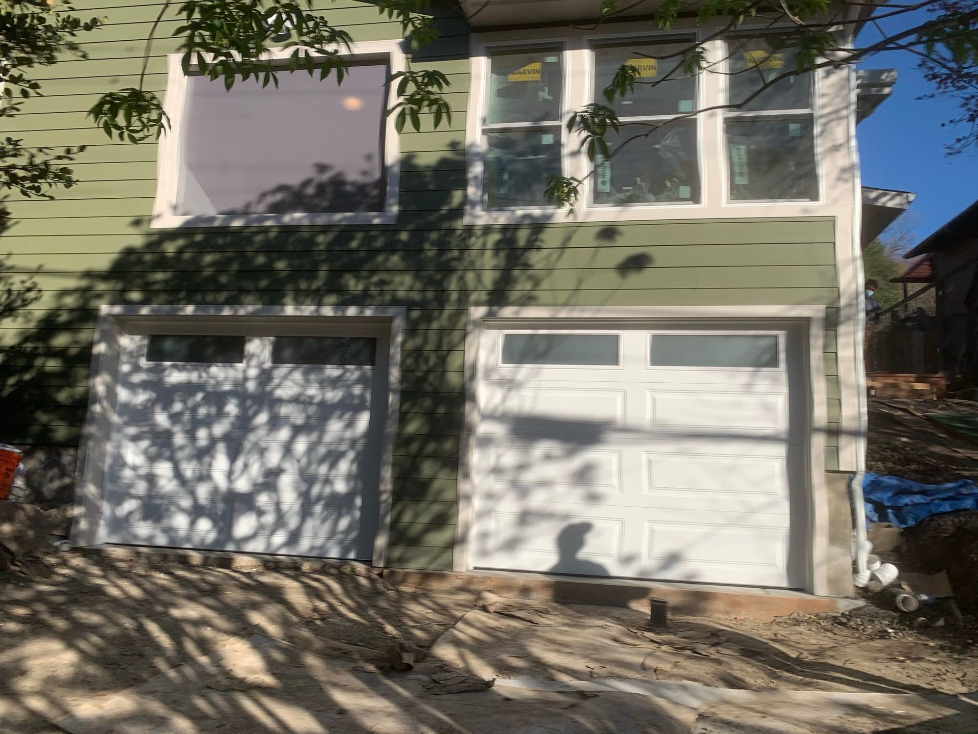 Two-car garage with white doors on a light green house, partly shaded by tree branches