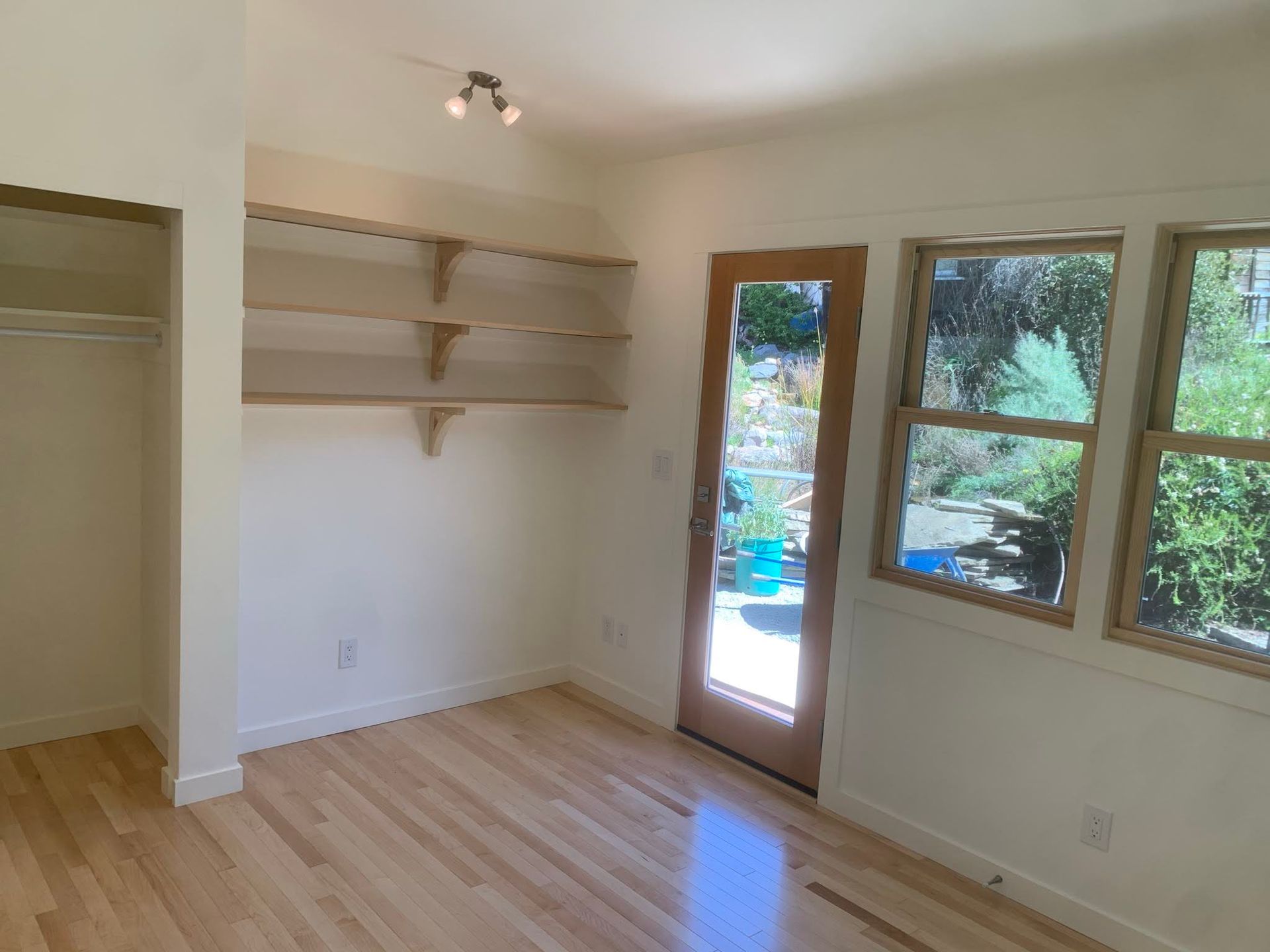 Empty beige room with light wood floor, wall shelves, closet, and windows by a glass door.