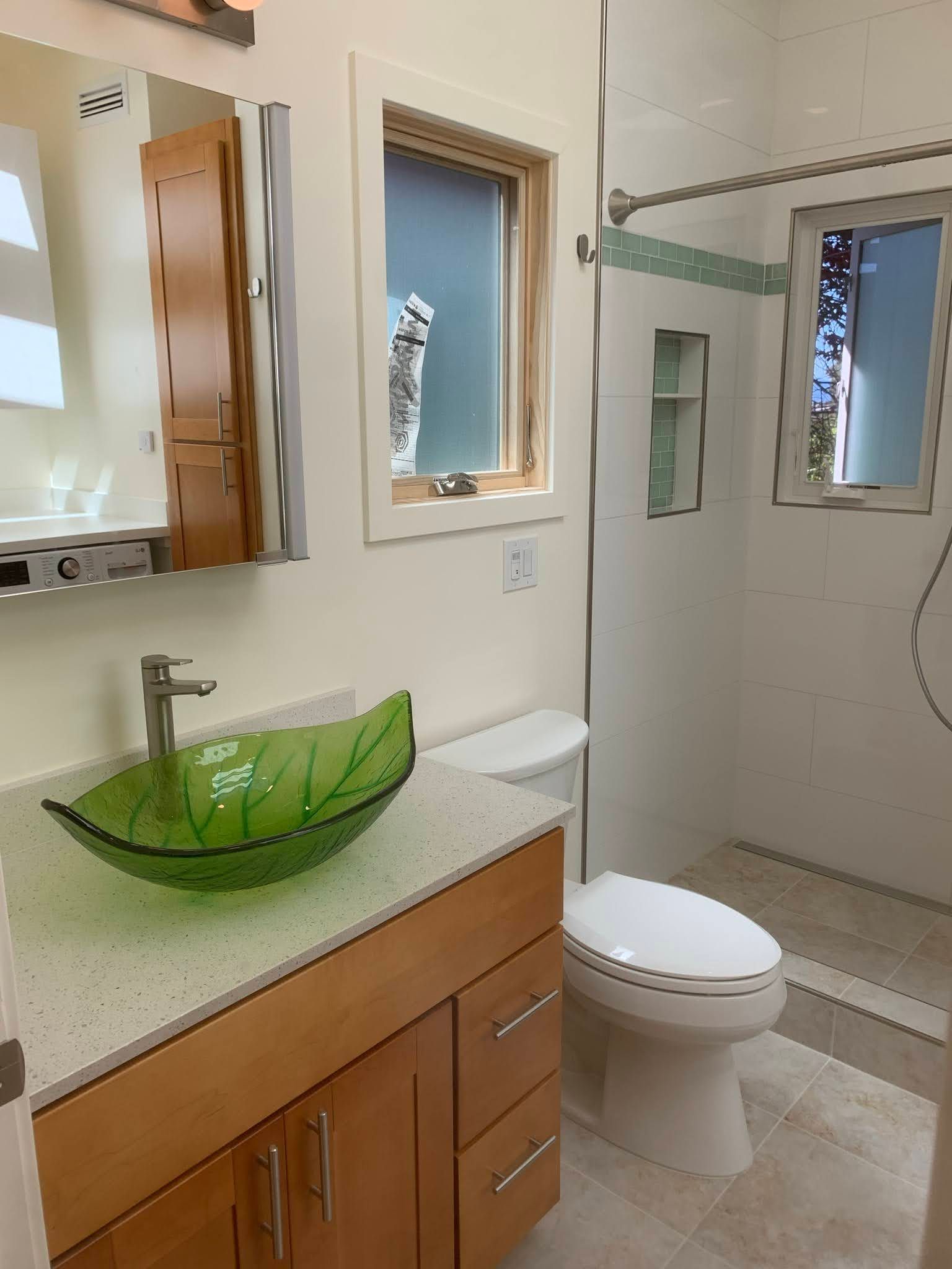 Modern bathroom with white toilet, green glass sink, wood vanity, mirror, and glass shower enclosure