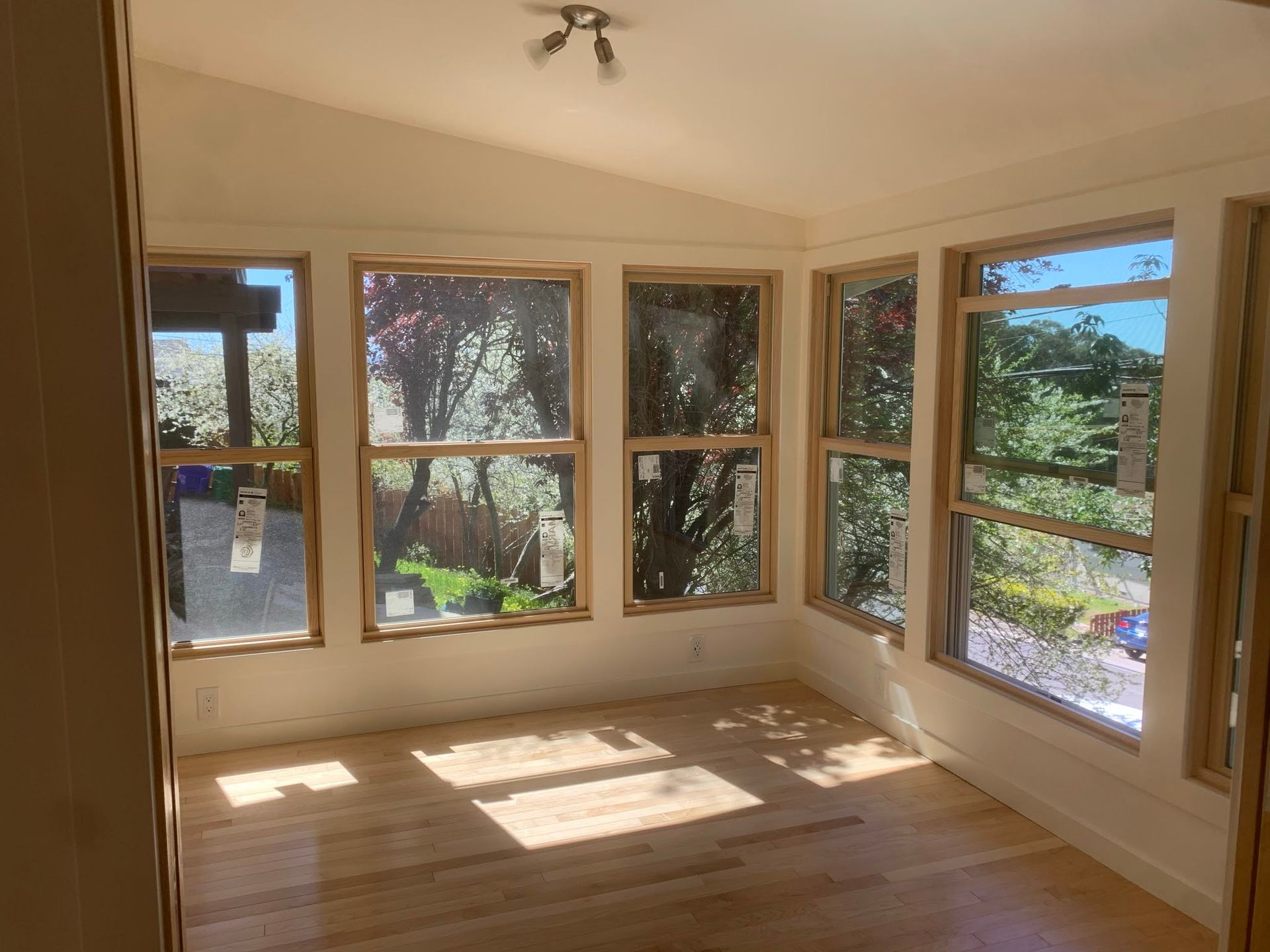 Empty sunlit room with large windows and hardwood floor, showing trees outside.