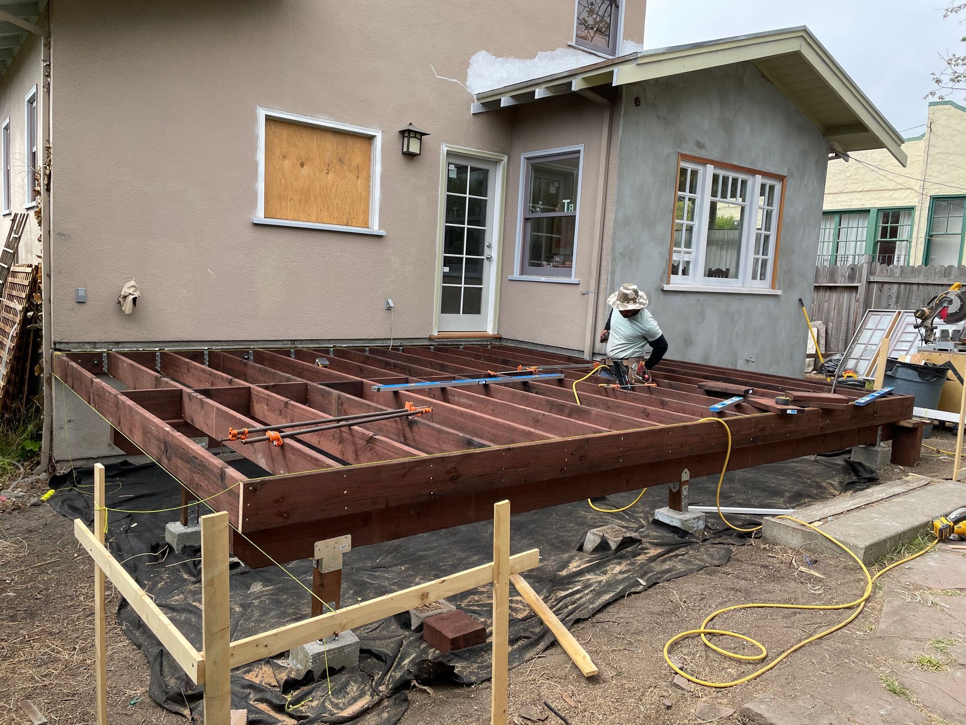 Worker building a raised wooden deck frame beside a house under construction.
