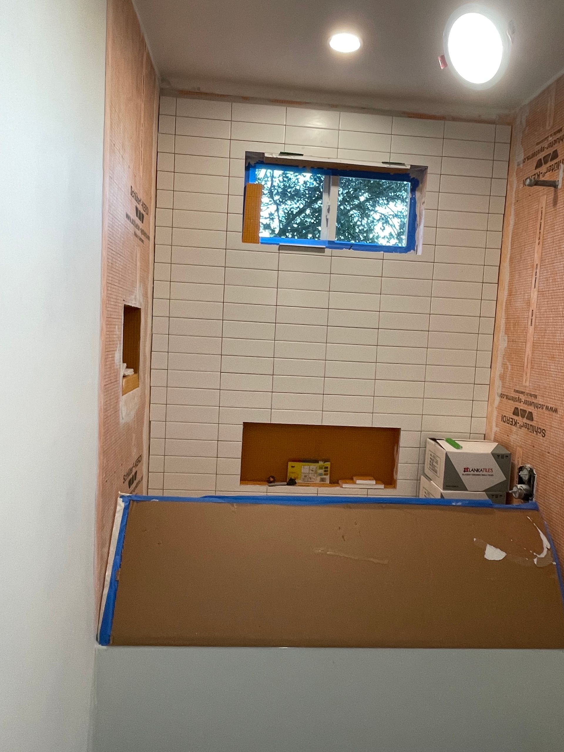 Small bathroom under renovation with tiled walls, a window, and a tub or shower covered by cardboard.