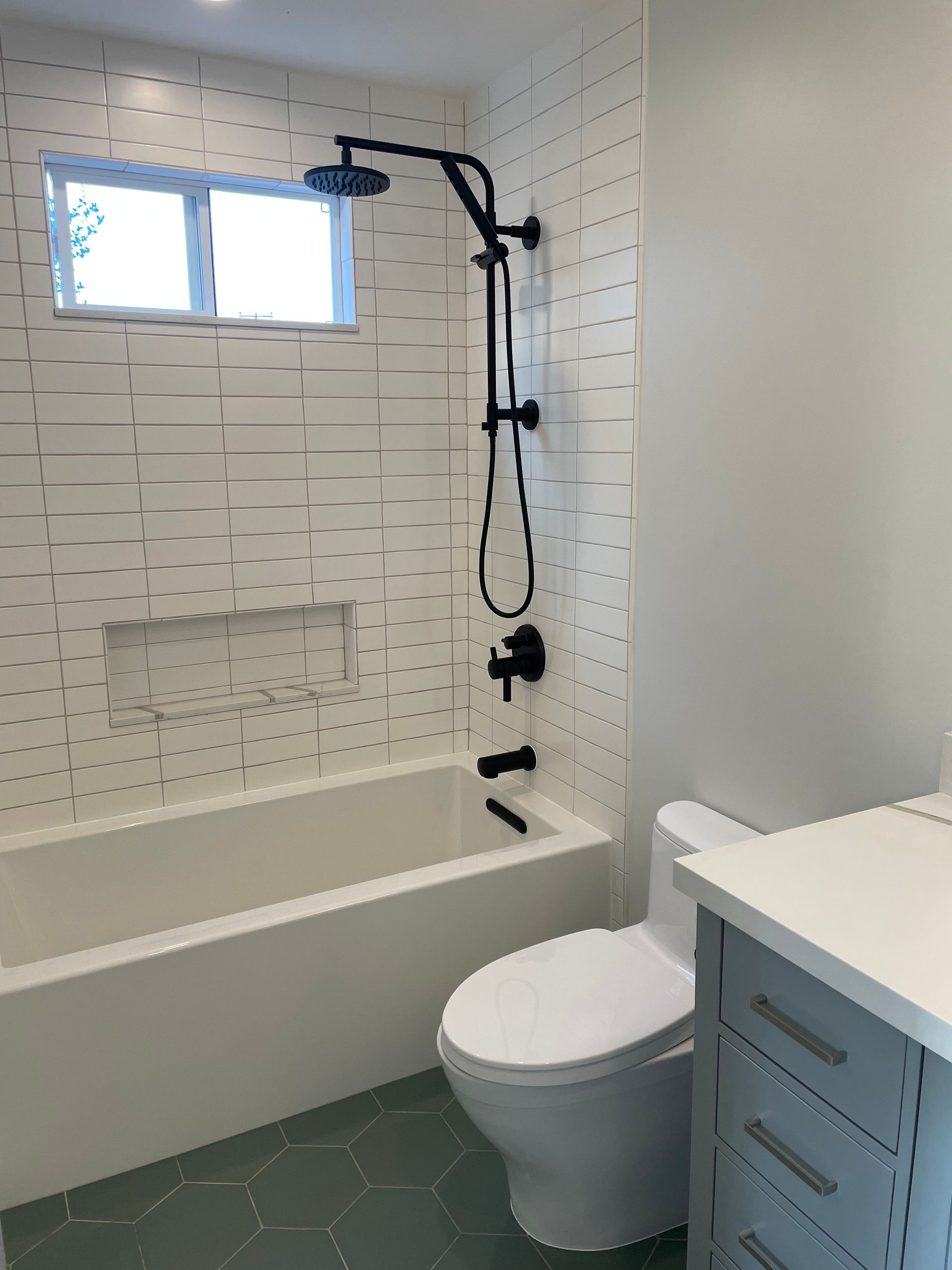 Small bathroom with bathtub, black shower fixture, white tile, and toilet beside a vanity.