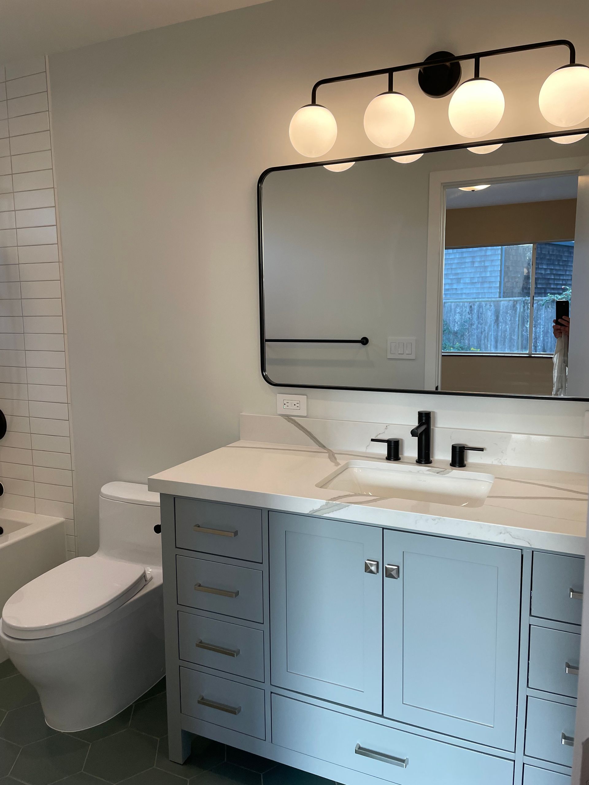 Modern bathroom with blue vanity, black fixtures, round mirror, and toilet beside a bathtub