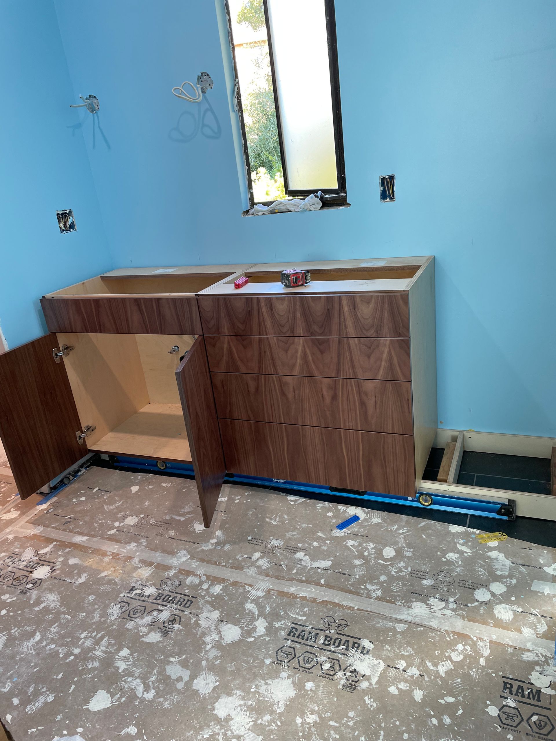 Kitchen cabinets being installed in a blue-walled room with a small window and unfinished floor.
